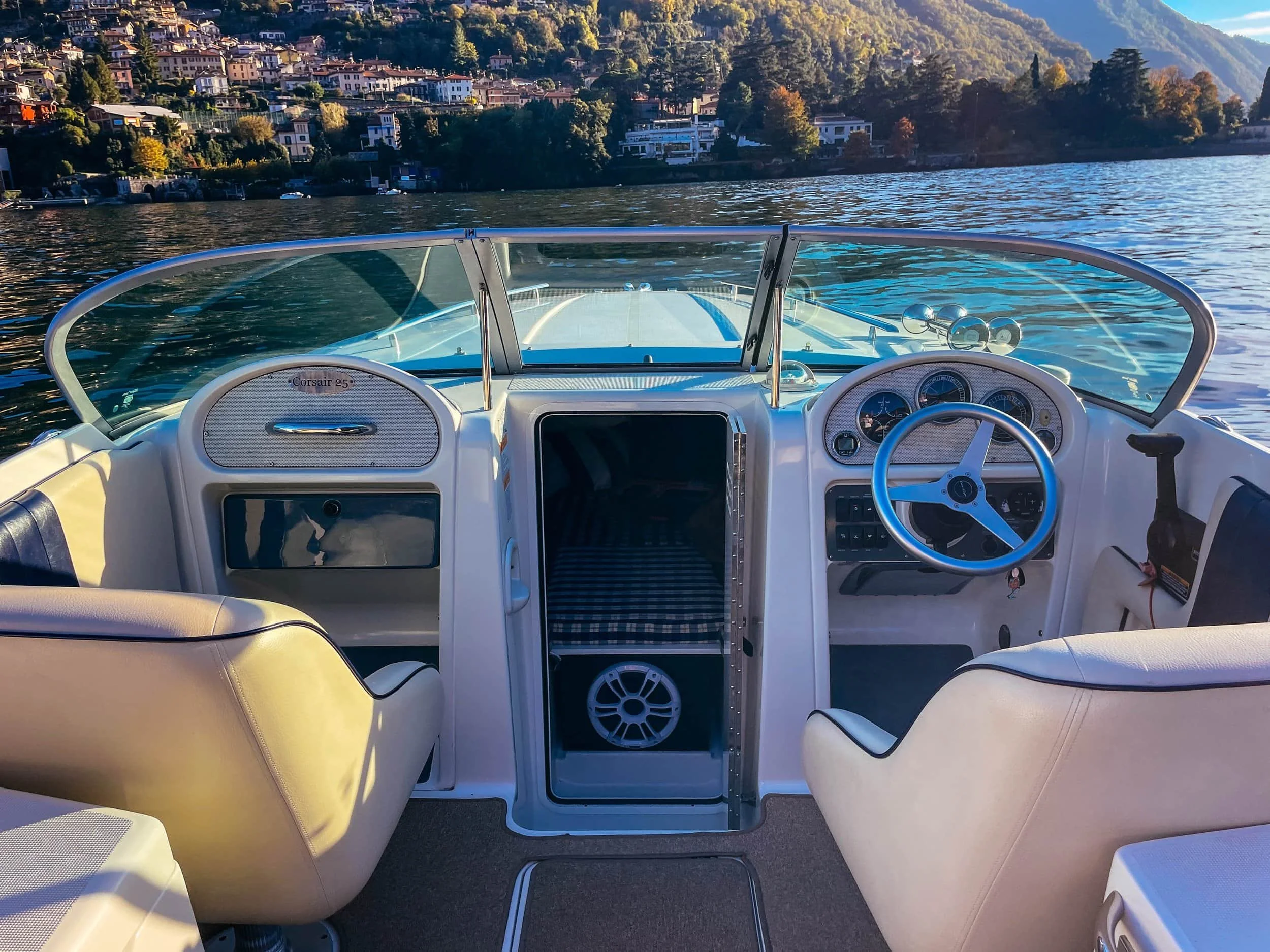 View of the cockpit of a Chris Craft speedboat with cream-colored seats, a steering wheel, and dashboard gauges, with a scenic Lake Como and surrounding hillside in the background.