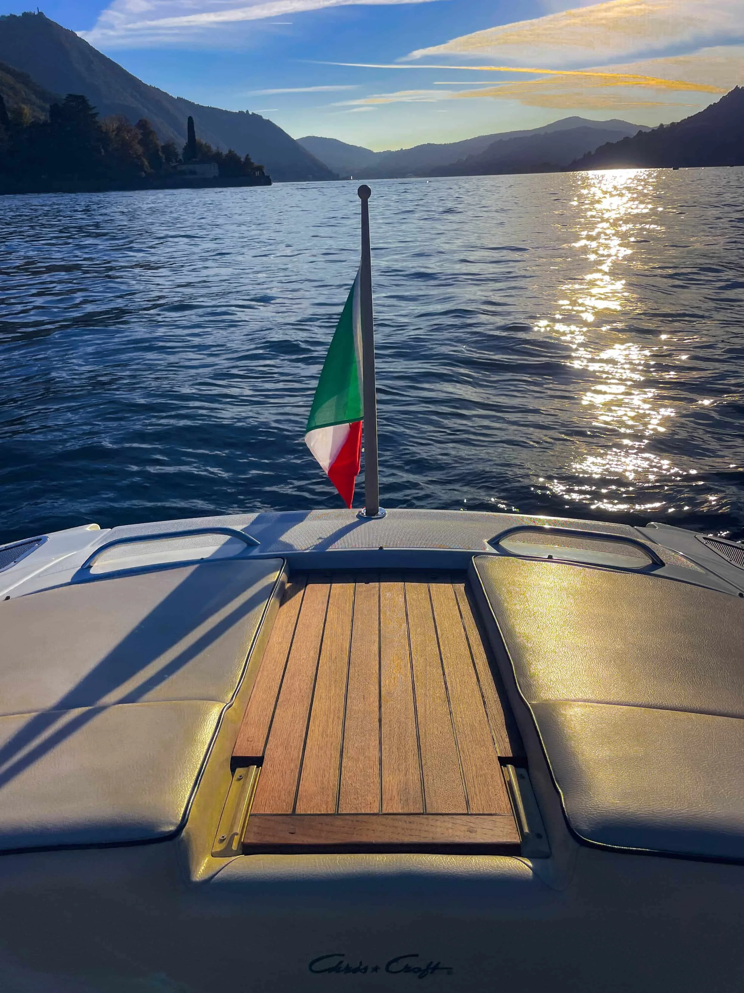 View from Chris Craft Corsair speedboat looking over Lake Como with Bellagio in the distance at sunset. An Italian flag is at the boat's stern. The boat's cushioned seating and wooden deck are visible in the foreground.