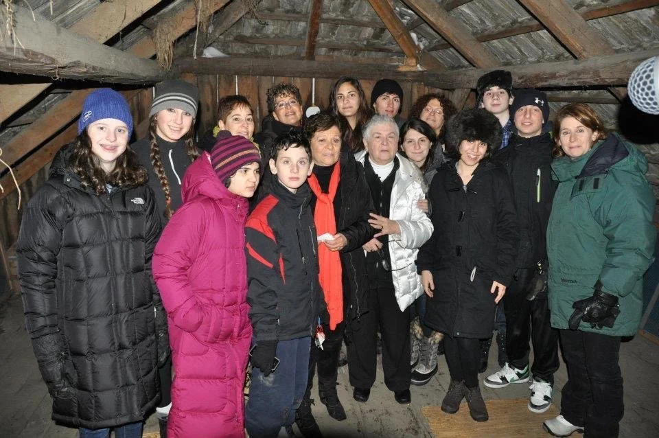 Sally Frischberg (front center, in white coat) and her sister Miriam (next to her, with red scarf) standing with their descendants in the attic where their family was hidden, 2012