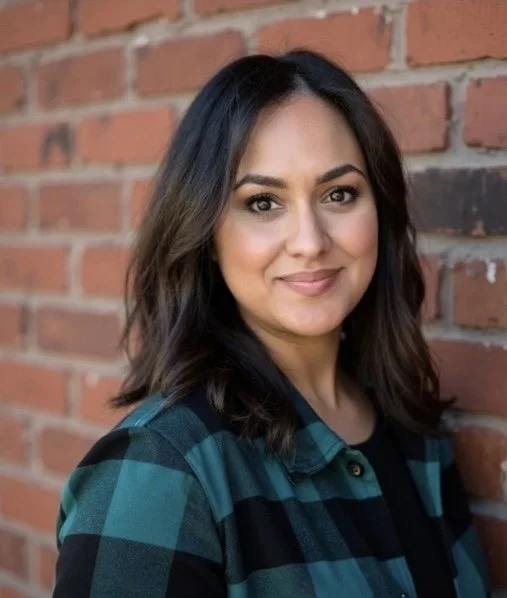 A woman with shoulder-length dark brown hair and light skin, smiling and looking at the camera, standing in front of a brick wall.