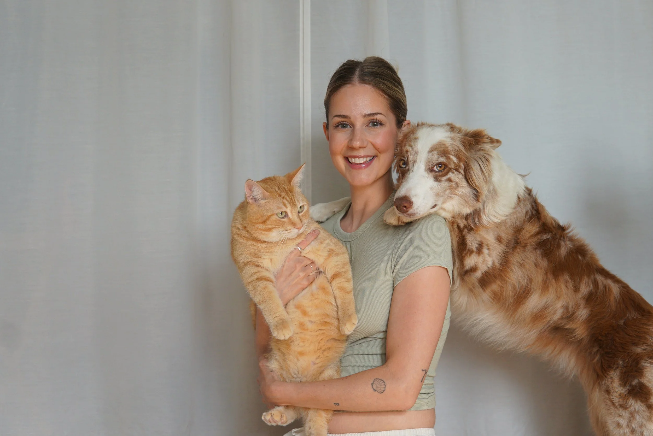Une femme vêtue d'un chandail vert sauge tien dans ses bras un chat roux qui regarde un chien appuyé sur les épaule de la femme. Le chien est brun et blanc. Tous pose devant une toile blanche.