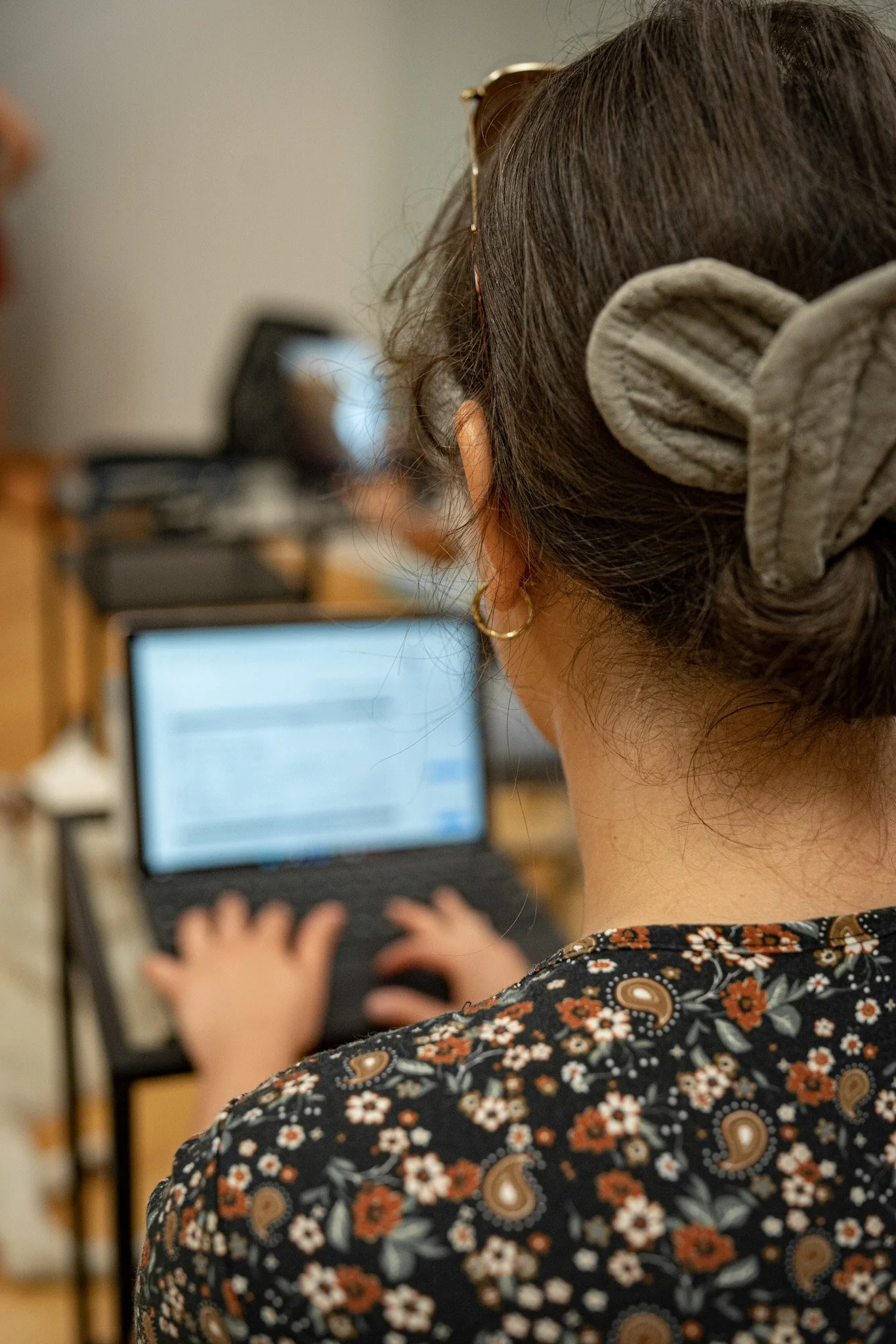 Une femme de dos aux cheveux attachés en toque et portant un chandail noir fleuri utilise le clavier de son ordinateur portable dans une classe