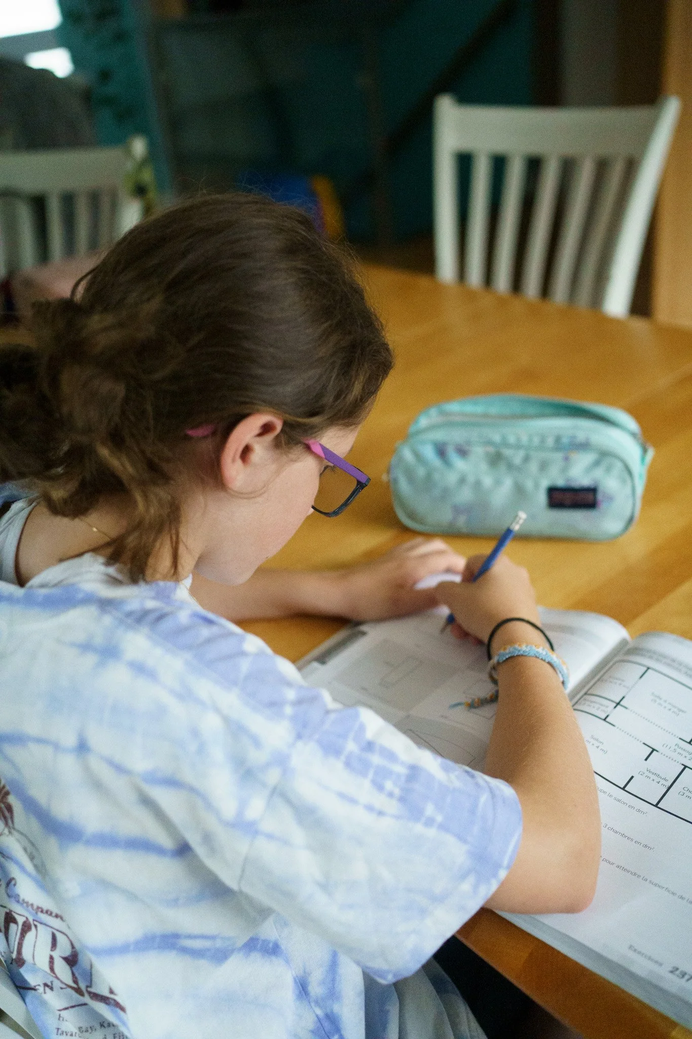 Une adolescente écrit dans un cahier assise à une table en bois. Elle porte des lunettes et un chandail blanc et mauve. Un coffre à crayon turquoise est placé tout près de son cahier