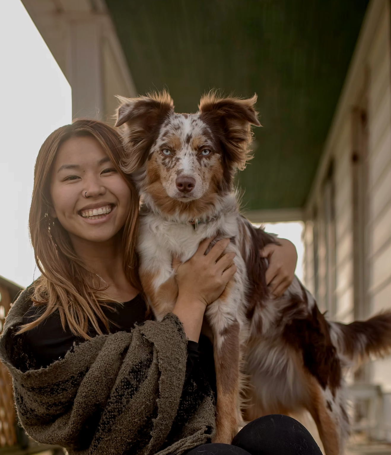 Une femme souriante aux cheveux longs est assise sur un porche, serrant dans ses bras un chien à la fourrure brune et blanche. La femme et le chien regardent tous deux la caméra, et l'arrière-plan montre une partie d'une maison.