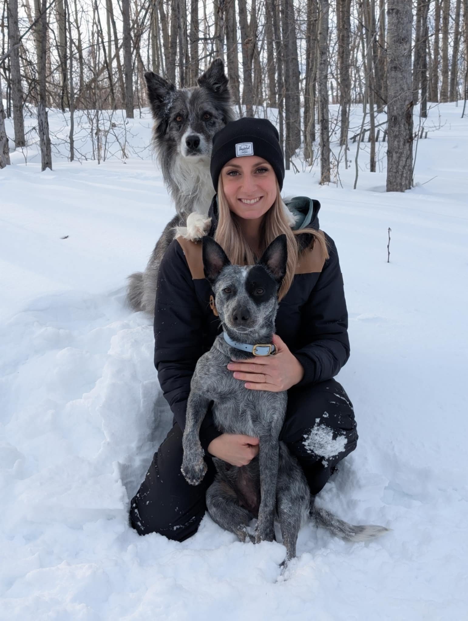 Une femme pose avec deux chiens : l'un est devant la femme et l'autre est derrière, avec 2 pattes sur les épaules de la femme. Ils sont dans une forêt enneigée.