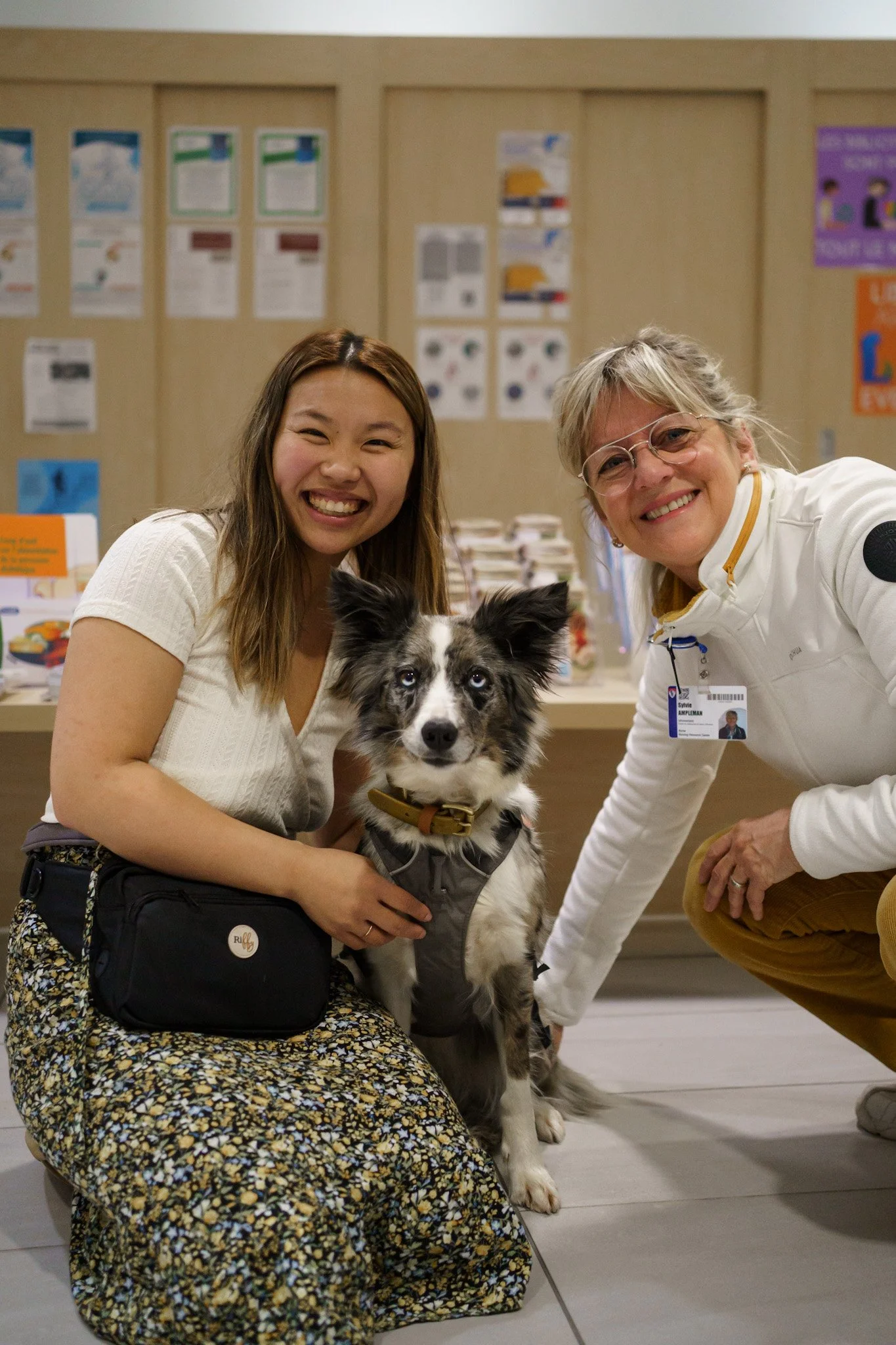 Deux femmes sourient à la caméra et entre elles, se trouvent un chien à la fourrures noire et blanche, portant un collier en cuir. Ils sont dans une salle d'événements avec une table derrière qui présente des affiches et pamphlets.