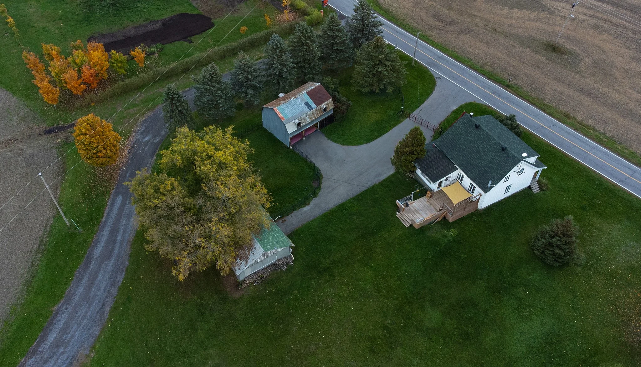 Vue aérienne d'une propriété rurale avec une maison blanche, une dépendance plus petite et une grange, entourée d'arbres, d'herbe verte et bordée d'une route et de feuillages d'automne.