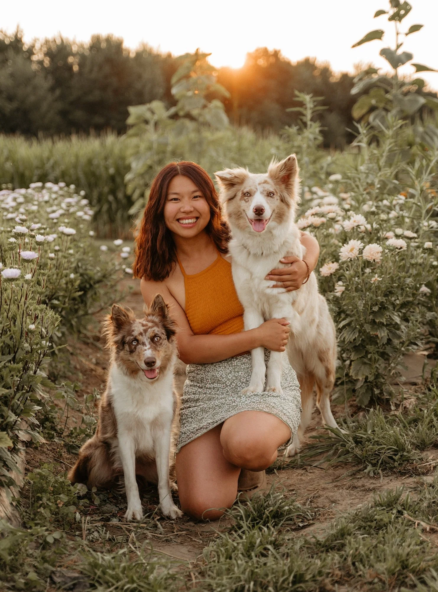 Une femme vêtue d'une camisole orange et d'une jupe aux motifs floraux pose dans un champs de fleurs avec deux chiens : l'un est assis par terre, l'autre est debout avec 2 pattes sur la femmes. Tous deux ont une fourrure brune et blanche.