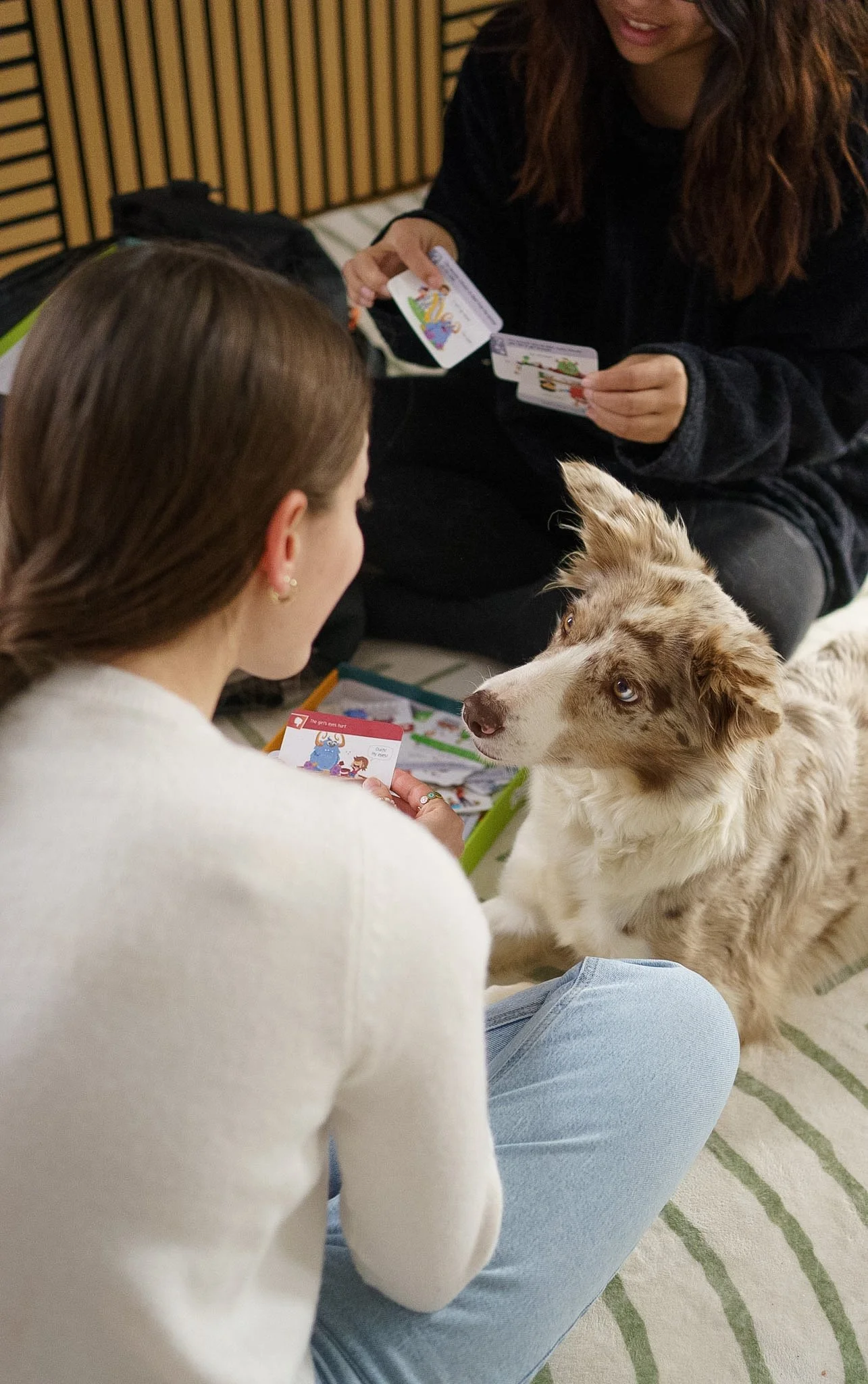 Deux femmes sont assises sur un tapis rayé et jouent à un jeu de société, avec un chien à côté d'elles. L'une d'elles sourit et brandit une carte de jeu, tandis que l'autre regarde le chien.