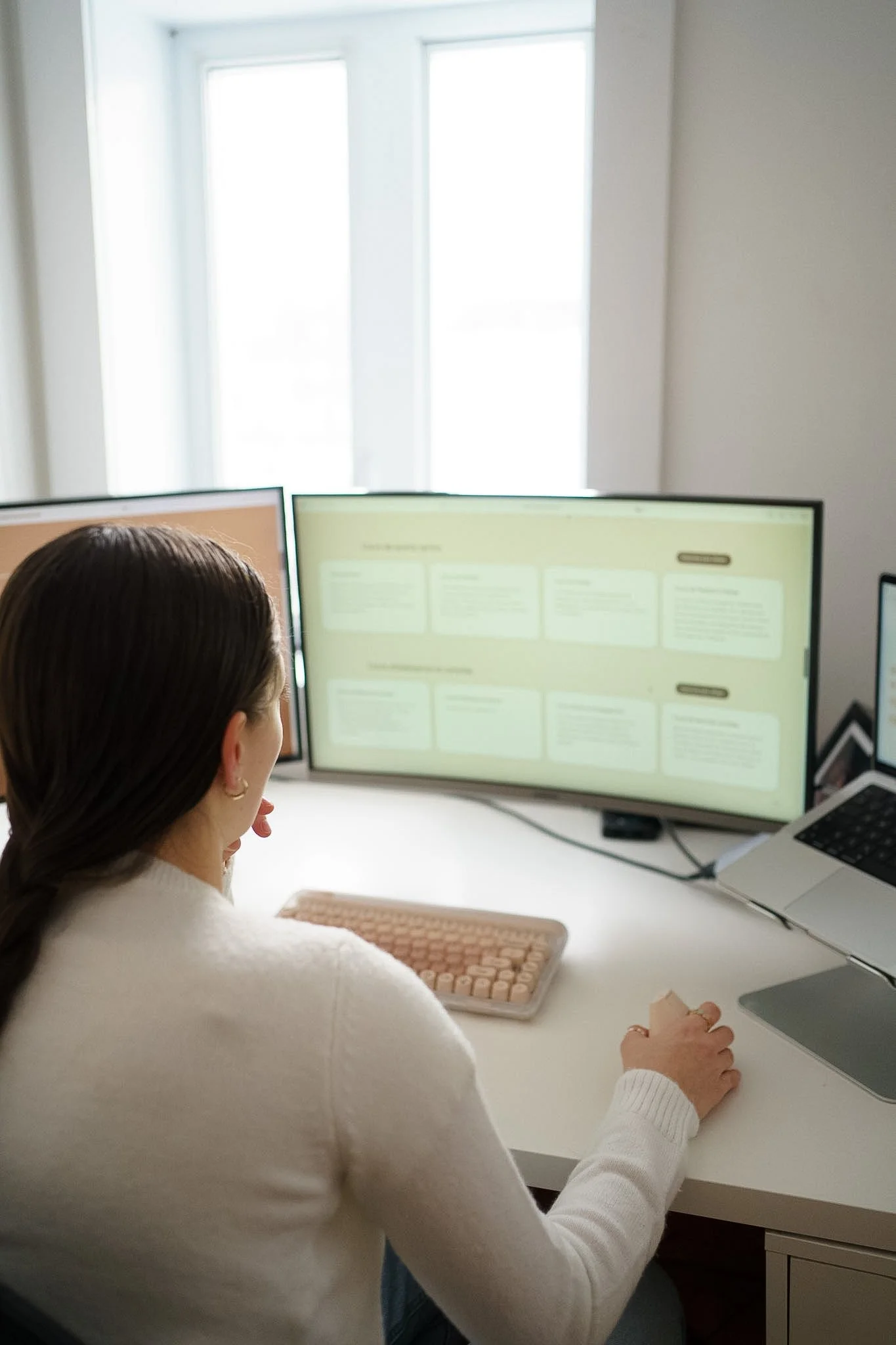 Une femme aux cheveux foncé et chandail blanc en laine navigue sur internet sur son ordinateur, un clavier et souris d'ordinateur rose