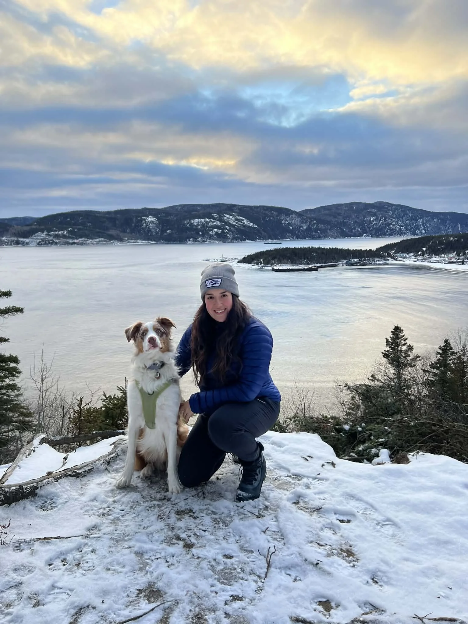 Une femme vêtue d'un manteau bleu et une tuque pose avec un chien blanc et brun devant un paysage montagneux et avec un grand lac lors d'une journée d'hiver.