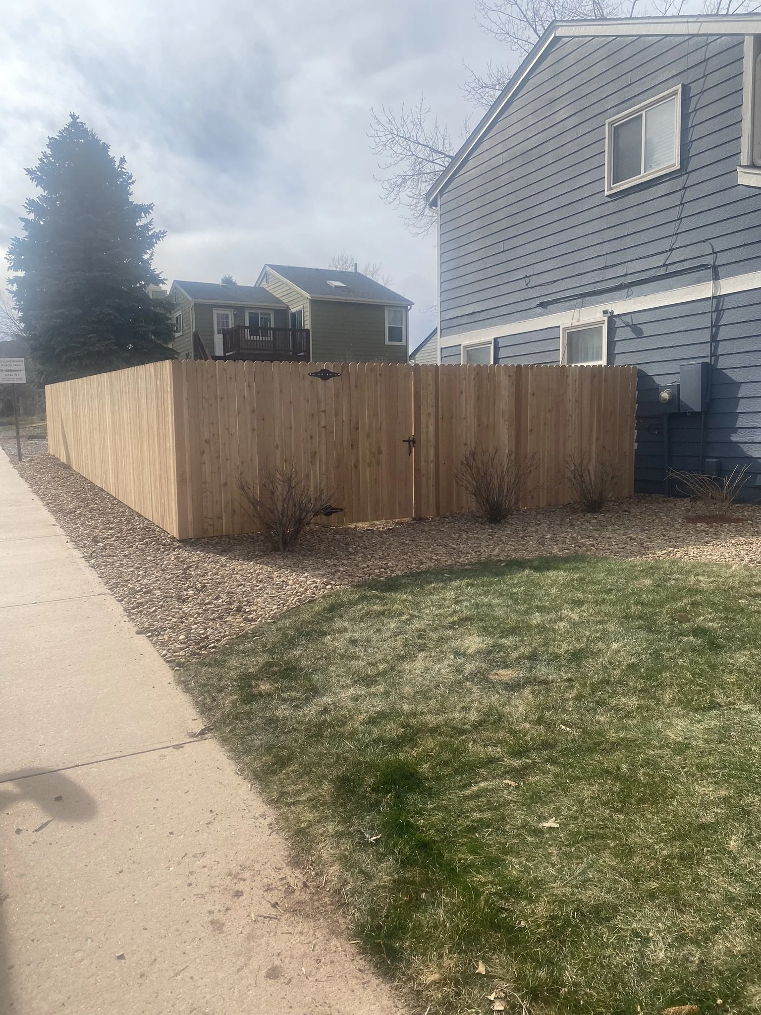 Photo of a backyard with a newly constructed wooden fence surrounding the yard, small bushes along the fence, a patch of grass in the foreground, and neighboring houses and a sidewalk in the background under a partly cloudy sky.