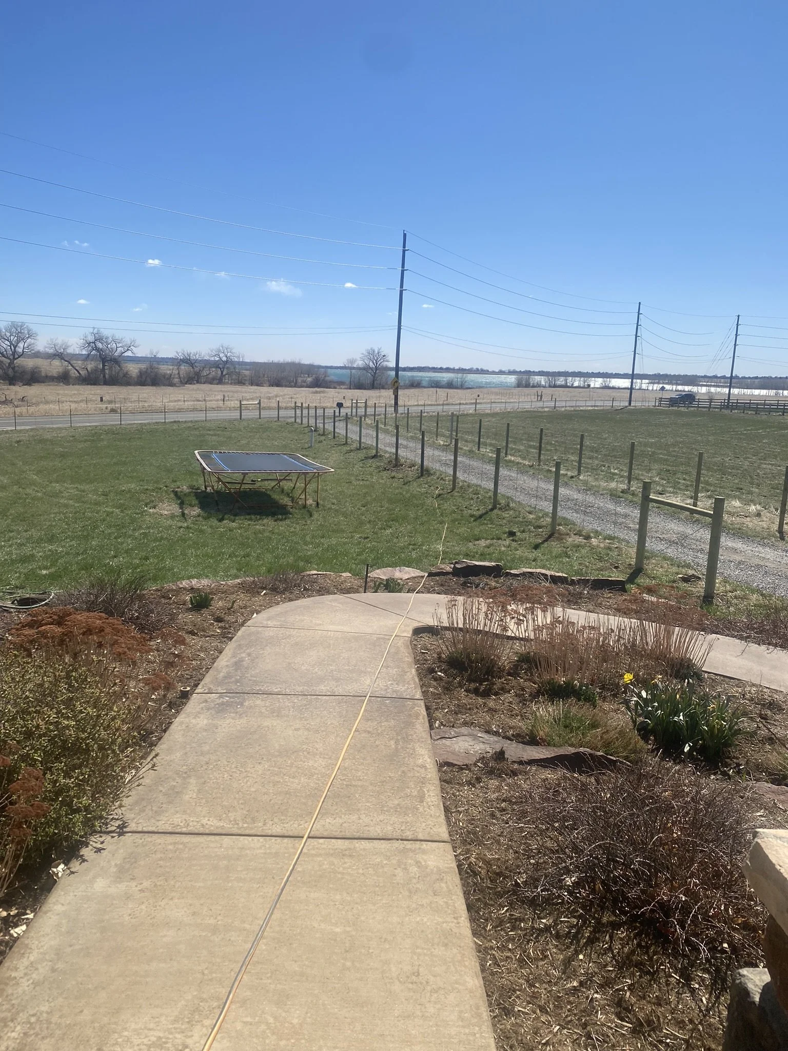 A backyard with a concrete sidewalk, small plants, and bushes, leading to a green yard enclosed by a wooden fence. Beyond the fence, there is a grassy field, a trampoline on the grass, and power lines with poles stretching across the sky. In the background, there are trees, open fields, and a blue sky with a few scattered clouds.