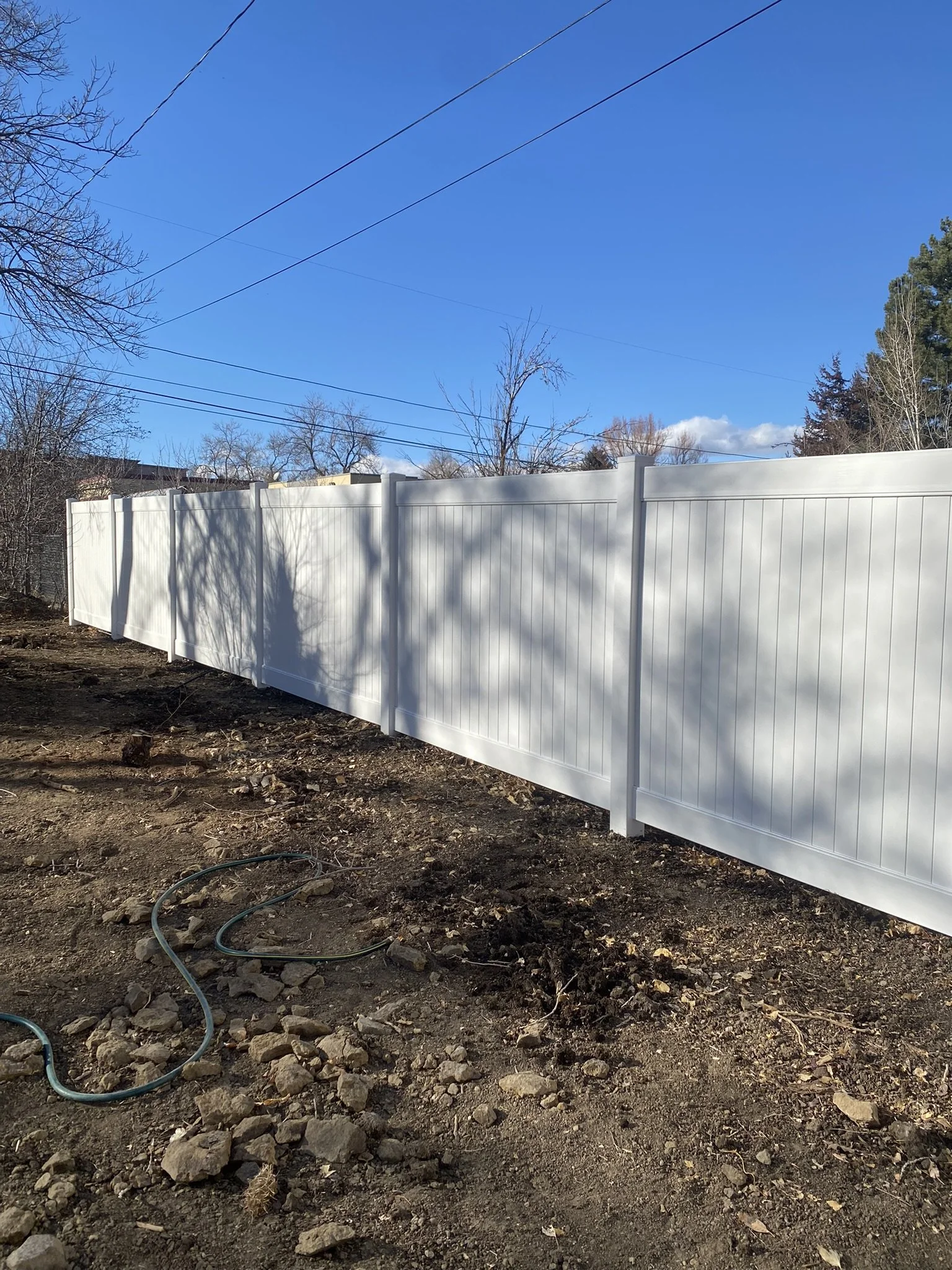 A white vinyl privacy fence installed along a dirt yard with a garden hose on the ground, under a clear blue sky with some trees in the background.