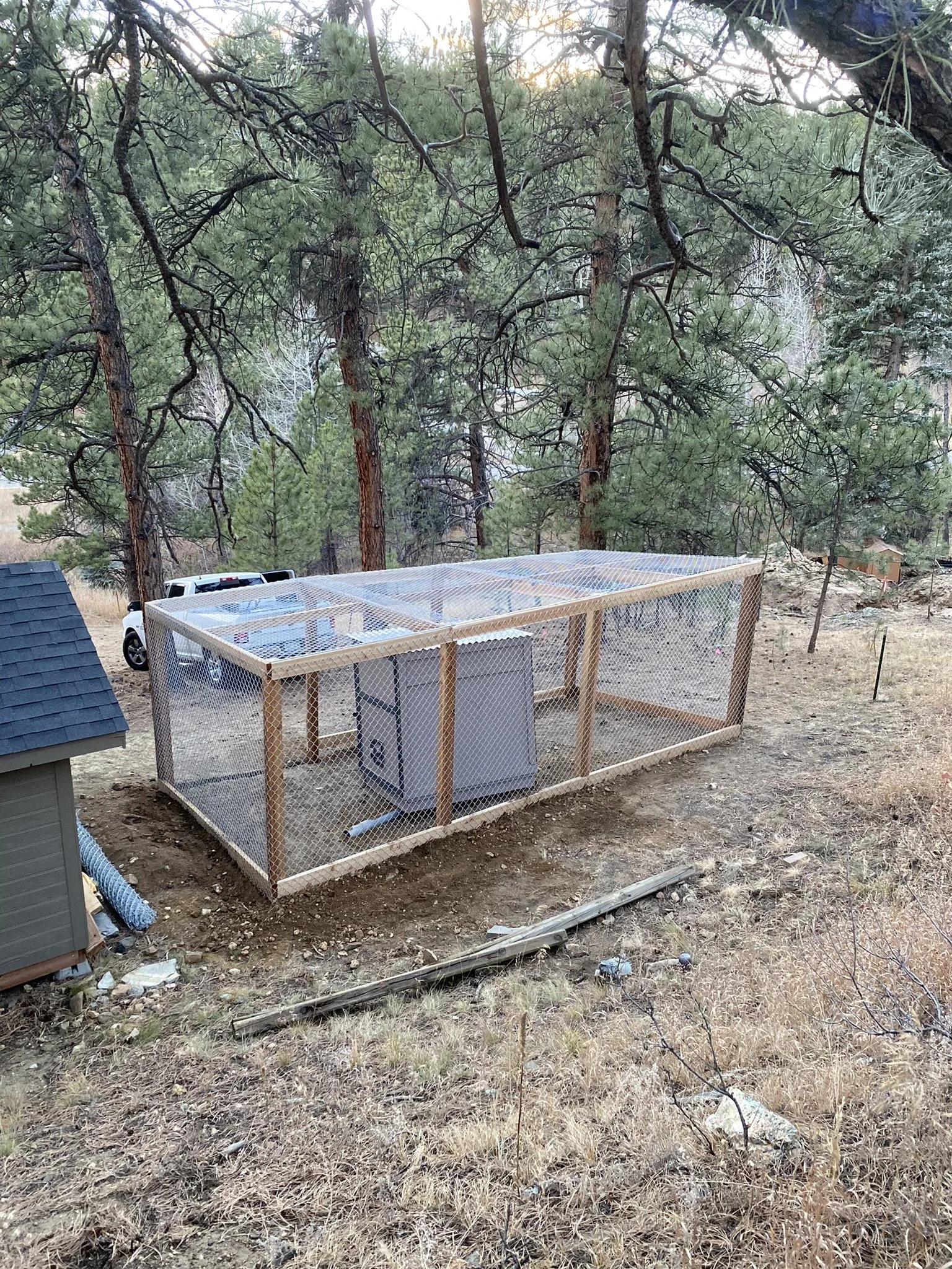 A fenced enclosure with a mesh wire exterior and a wooden frame, containing a white appliance and some piping, situated on a dirt patch surrounded by dry grass with a large pine tree in the background, and a vehicle partially visible behind the enclosure.