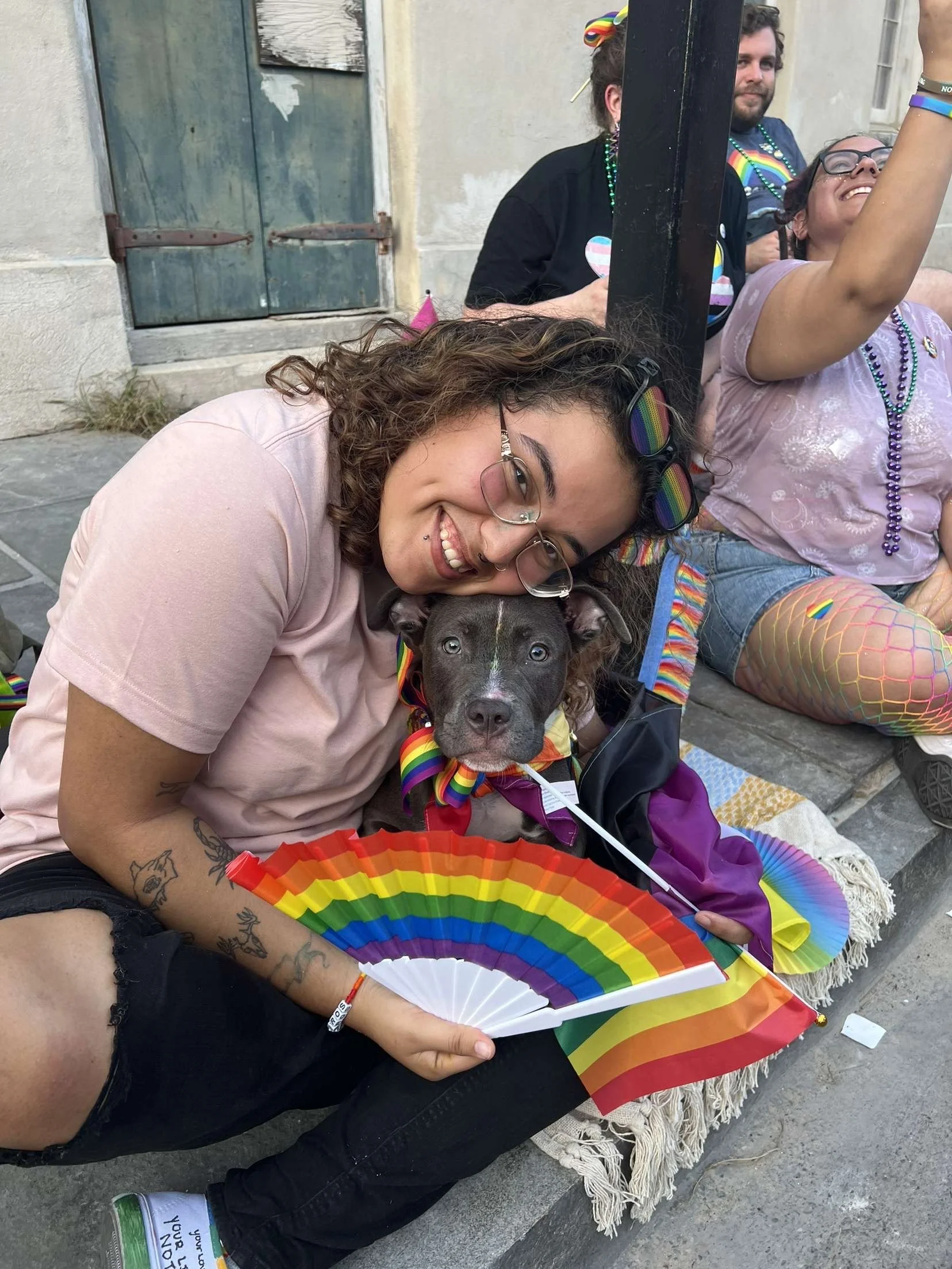 A group of people at a LGBTQ+ pride event, wearing rainbow accessories. In the foreground, a woman with curly hair, glasses, and tattoos is smiling as she leans over a dog, which is also wearing rainbow accessories. The woman is holding a rainbow fan, and others in the background are sitting on steps, smiling and wearing rainbow-themed clothing and jewelry.