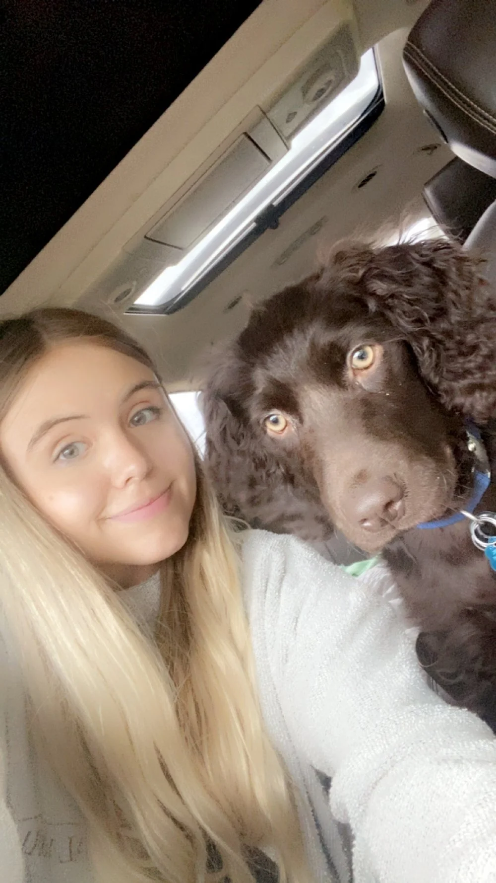A young woman with long blonde hair and light skin taking a selfie inside a vehicle, with a large brown dog with long ears and light eyes beside her.