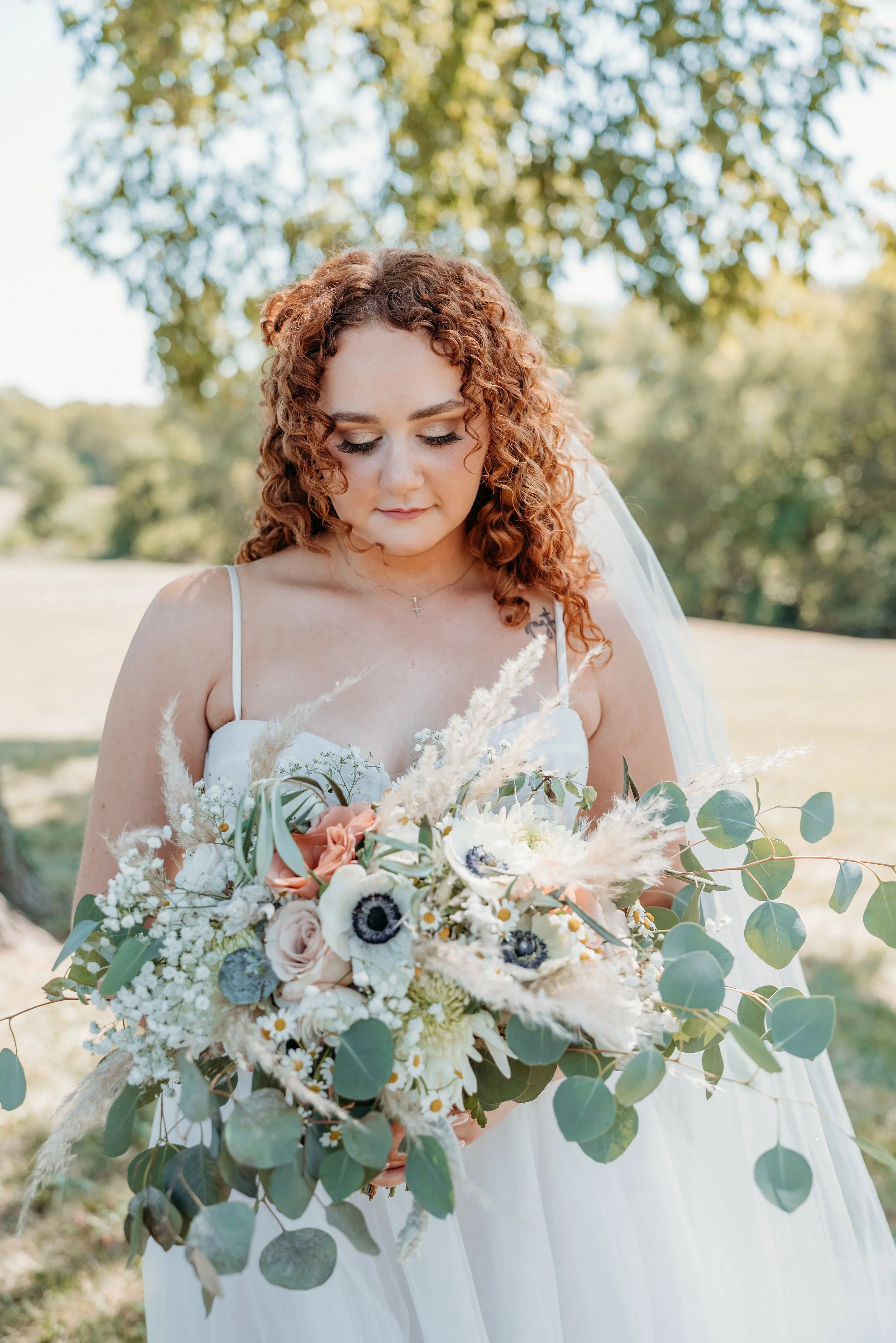 Bridal woman in white dress holding a bouquet of white, pink, and purple flowers with greenery, outdoors on a sunny day.