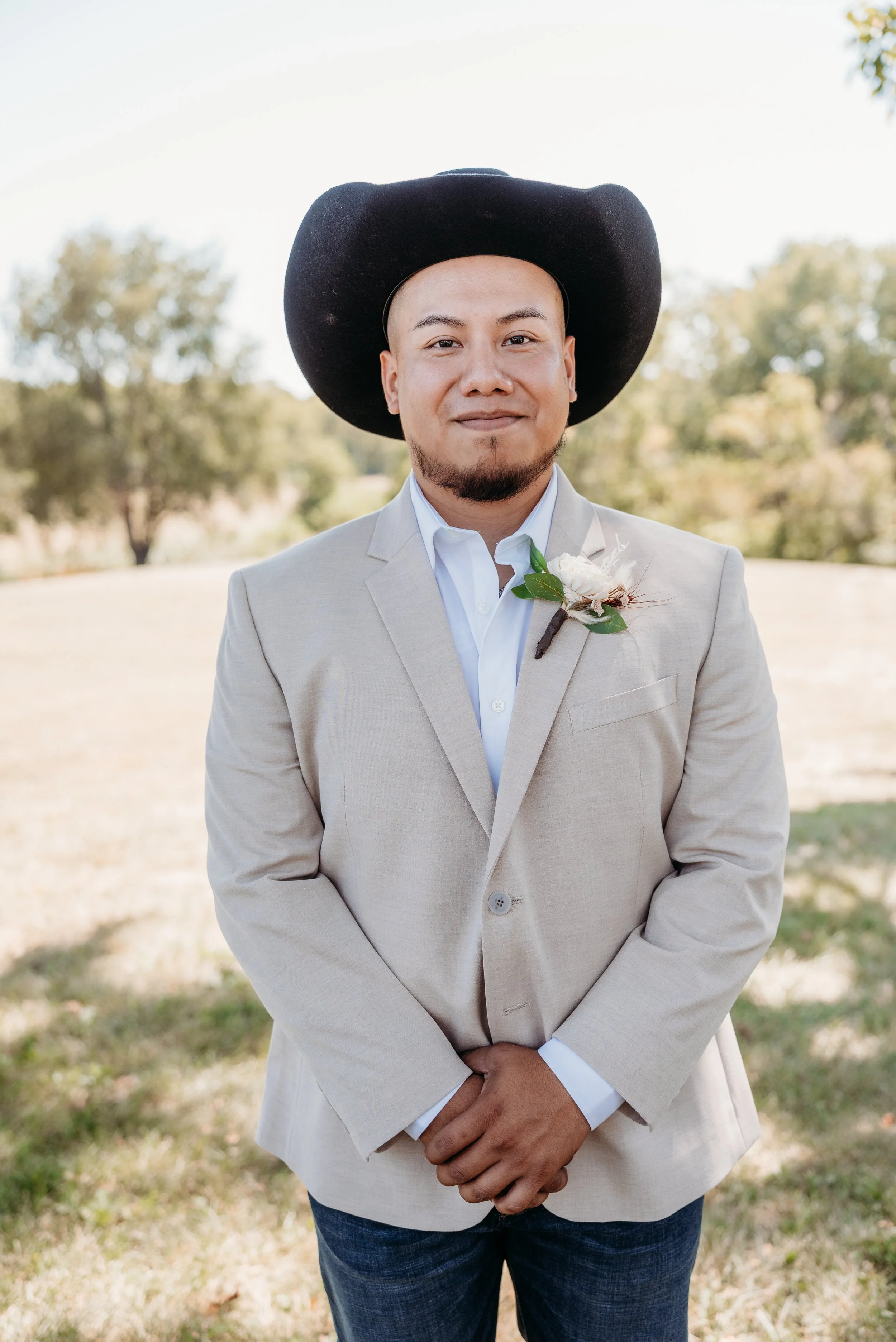 A man in a beige suit, white shirt, and a large black cowboy hat standing outdoors with trees in the background.
