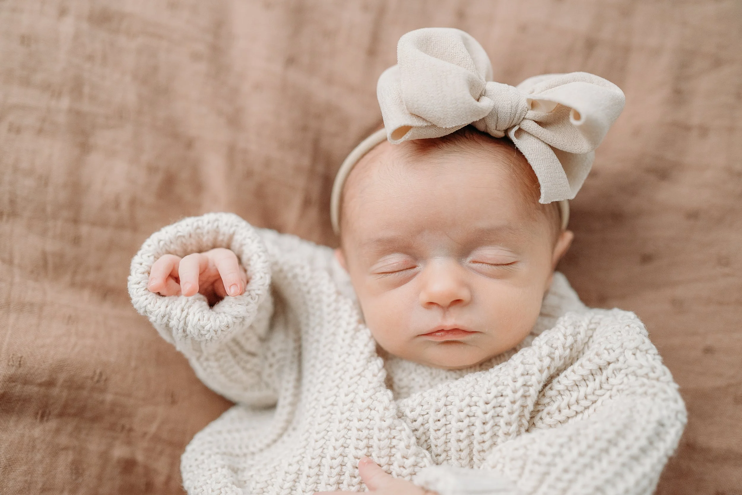 A sleeping baby girl wearing a beige bow headband and a knitted cream-colored sweater, lying on a brown fabric background.