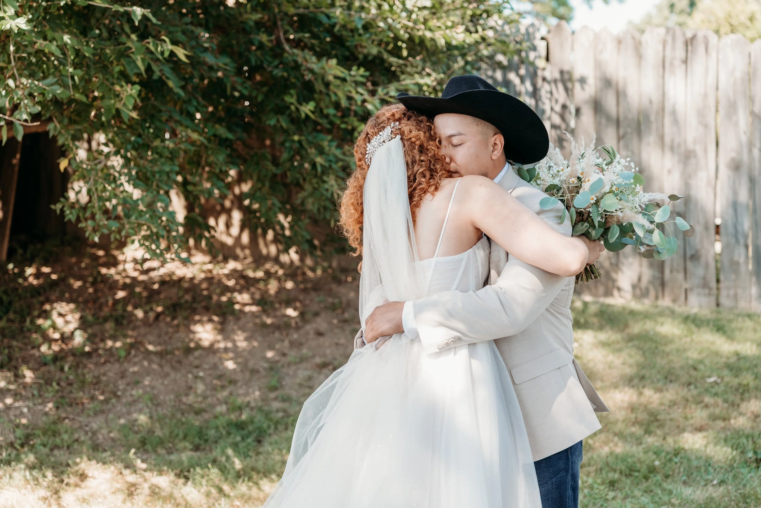 A bride and groom embracing outdoors, the bride in a white wedding dress with a veil and the groom in a beige suit and cowboy hat, holding a bouquet of flowers.