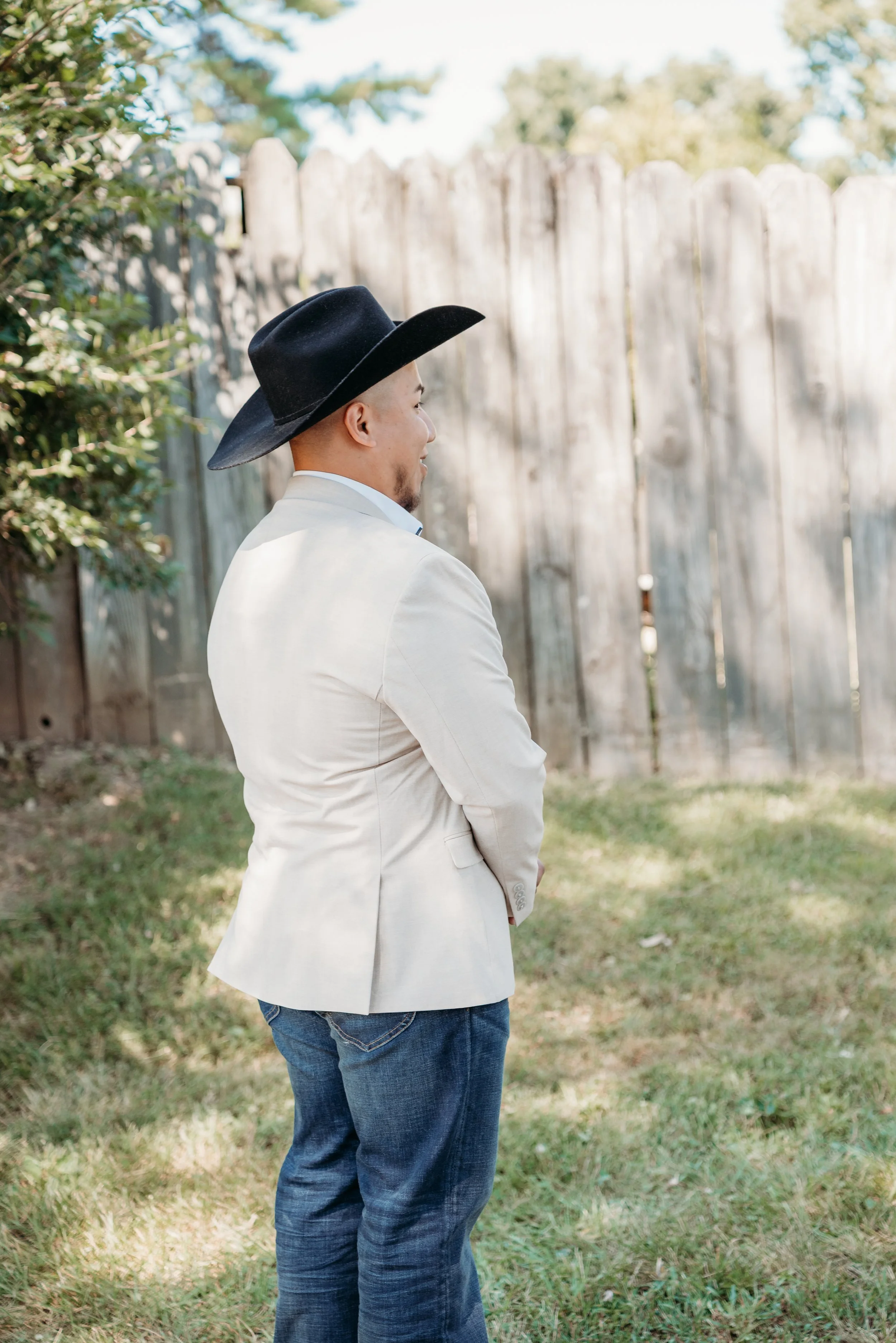 Man wearing a black cowboy hat, white blazer, and jeans standing in a backyard with a wooden fence, green grass, and trees.