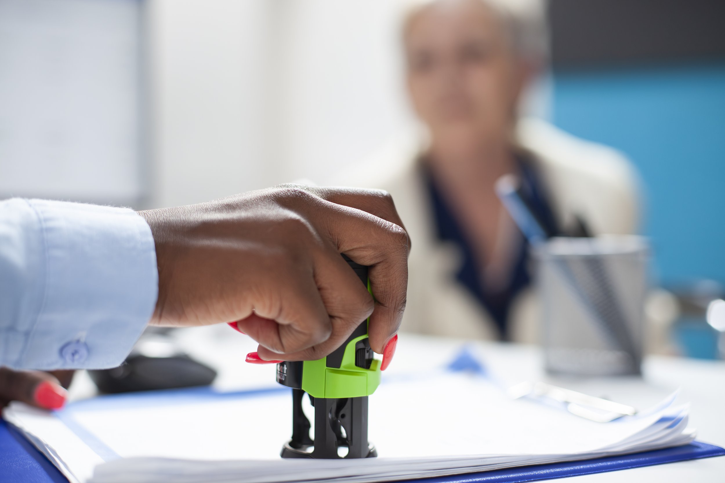 Close-up of a person's hand pressing a green button on a device on a stack of papers, with a blurred woman in a white blazer and dark shirt sitting in the background at a desk.