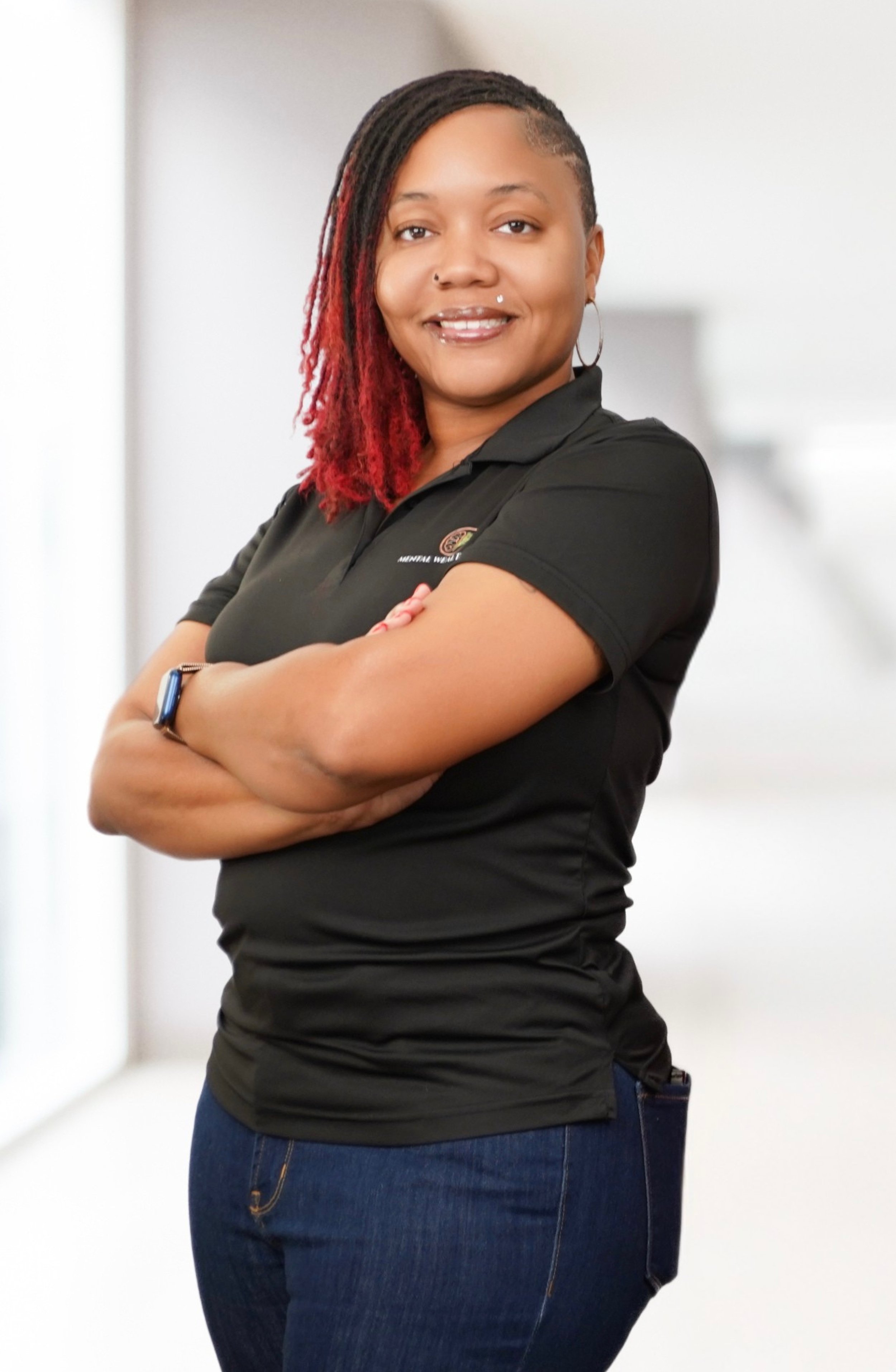 A young woman with long dark hair wearing a blue blouse, sitting in front of a neutral background with a floor lamp and a green leafy plant.