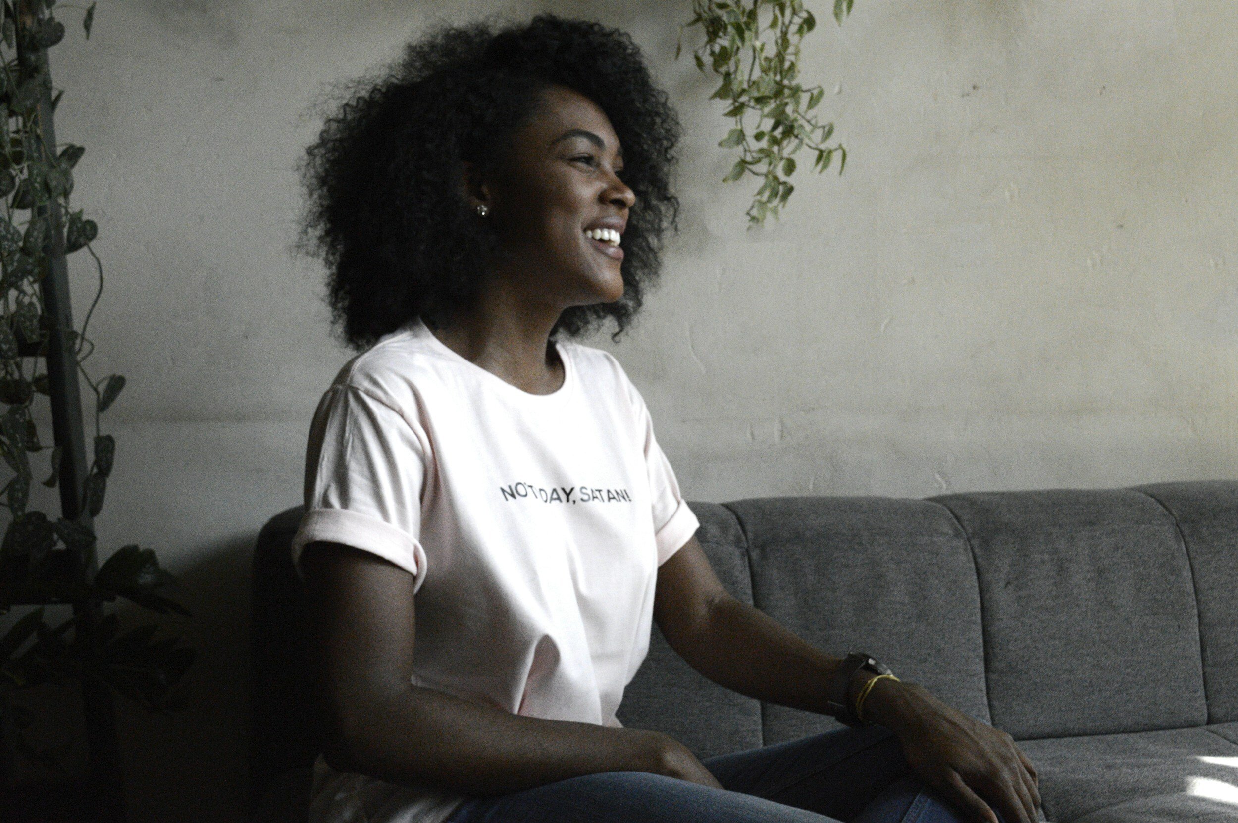 A woman with curly black hair smiling, sitting on a gray couch with a white brick wall and some greenery in the background. She is wearing a light pink t-shirt with the text "NOT TODAY, SATAN!" on it.