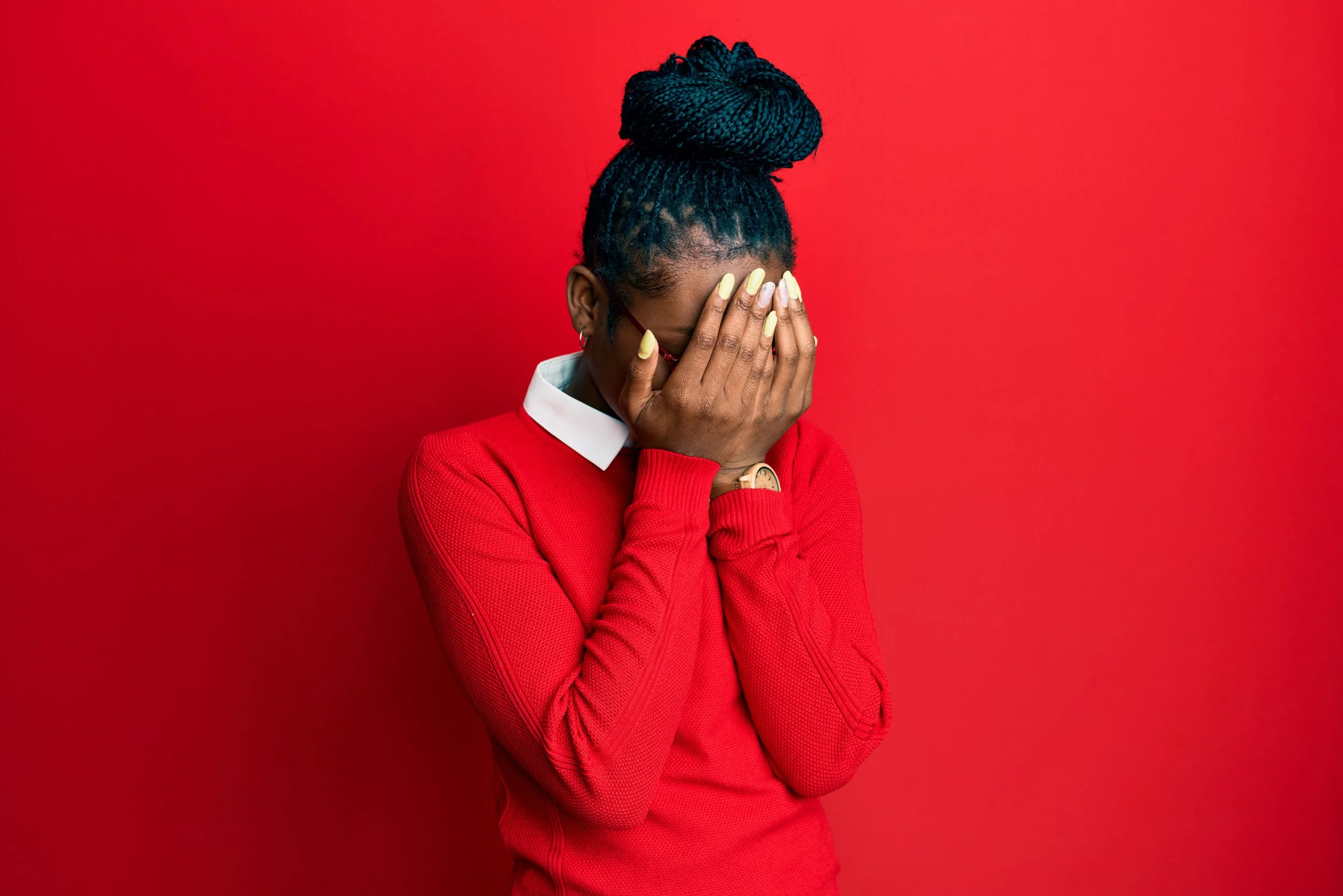 A woman with braided hair, wearing glasses and a red long-sleeve shirt with a white collar, covers her face with her hands, standing against a solid red background.