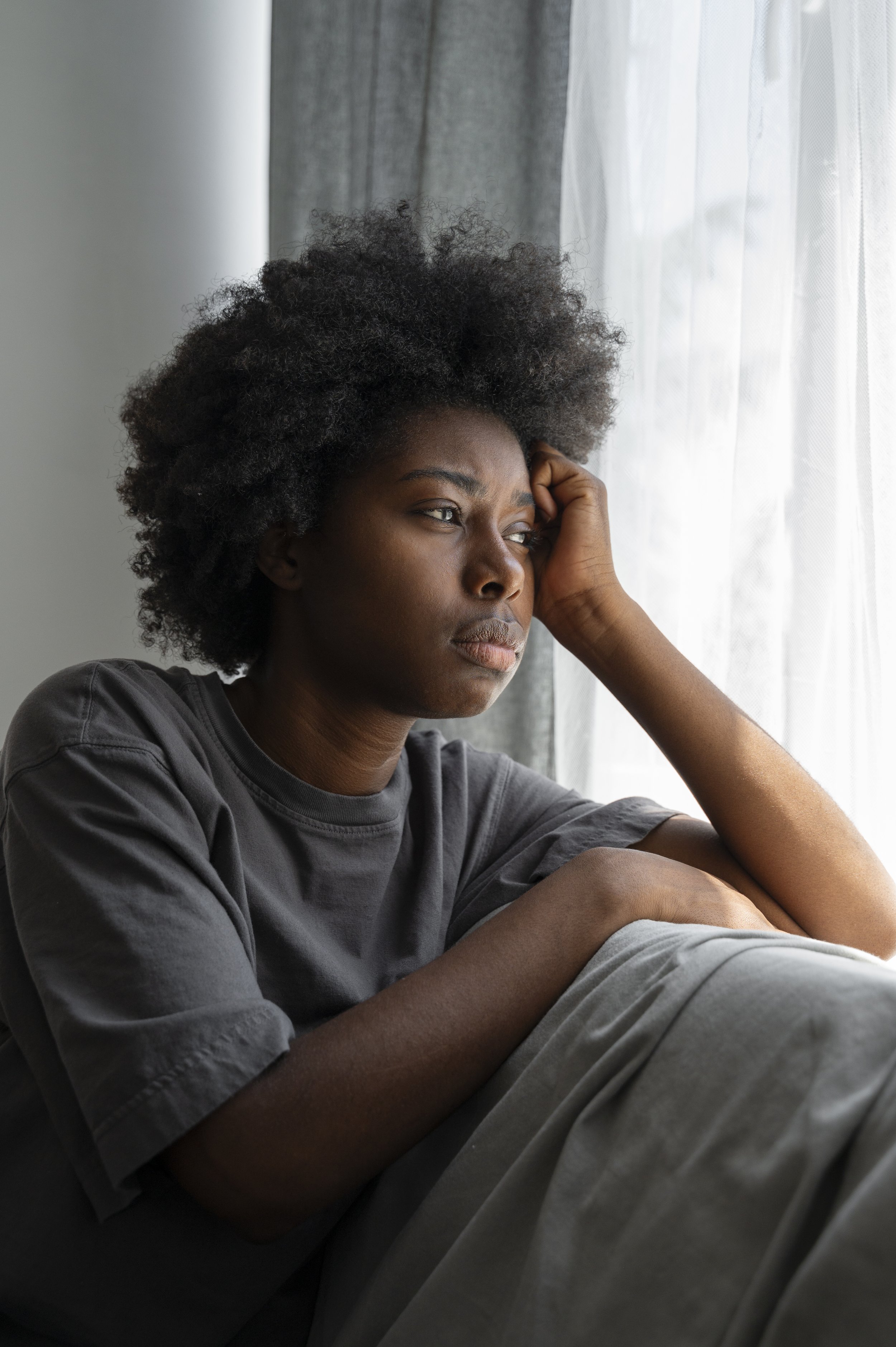 A woman with dark skin and short curly hair sitting by a window, looking out thoughtfully with a serious expression.