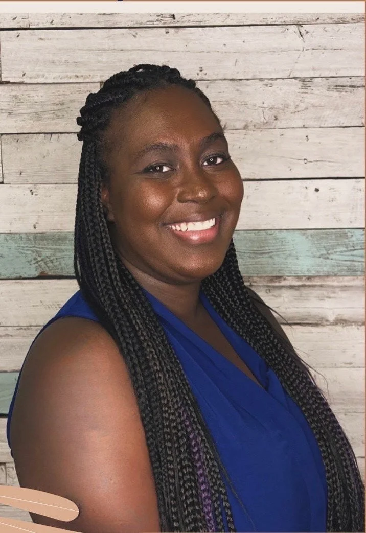 A smiling woman with braided hair wearing a blue sleeveless top, standing in front of a wooden panel wall.
