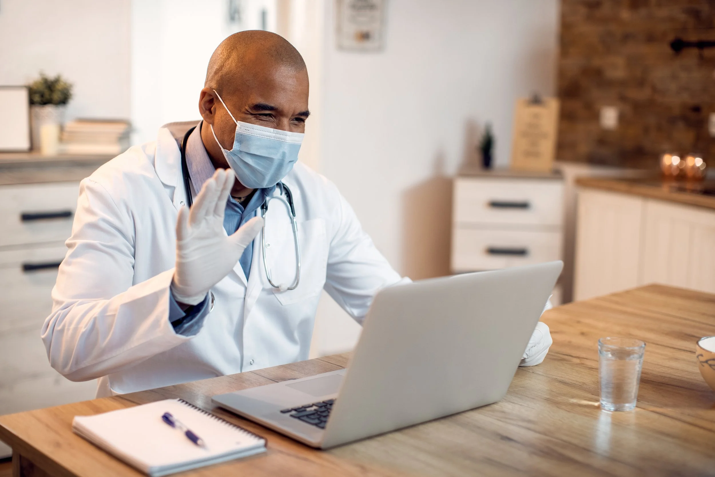 A doctor wearing a face mask, gloves, and a white coat is sitting at a wooden desk, waving at a video call on a laptop, with a notepad and pen in front of him and a glass of water nearby.