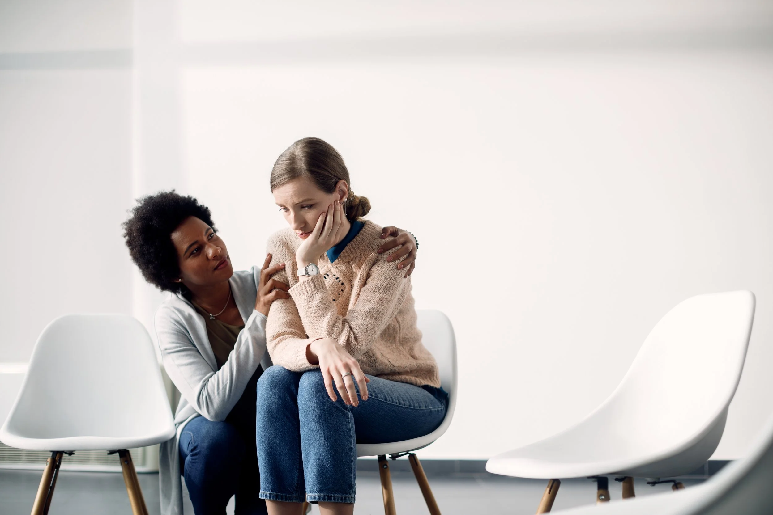 A woman appears distressed while sitting on a chair, comforted by another woman who is touching her shoulders. They are in a room with white walls and empty white chairs.