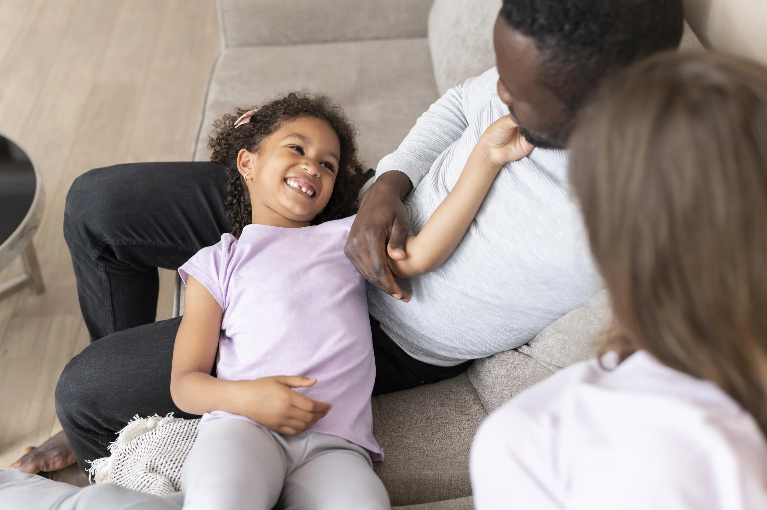 A girl is lying on her father's lap, smiling and playing with his arm, while her mother is sitting nearby, all on a beige couch.
