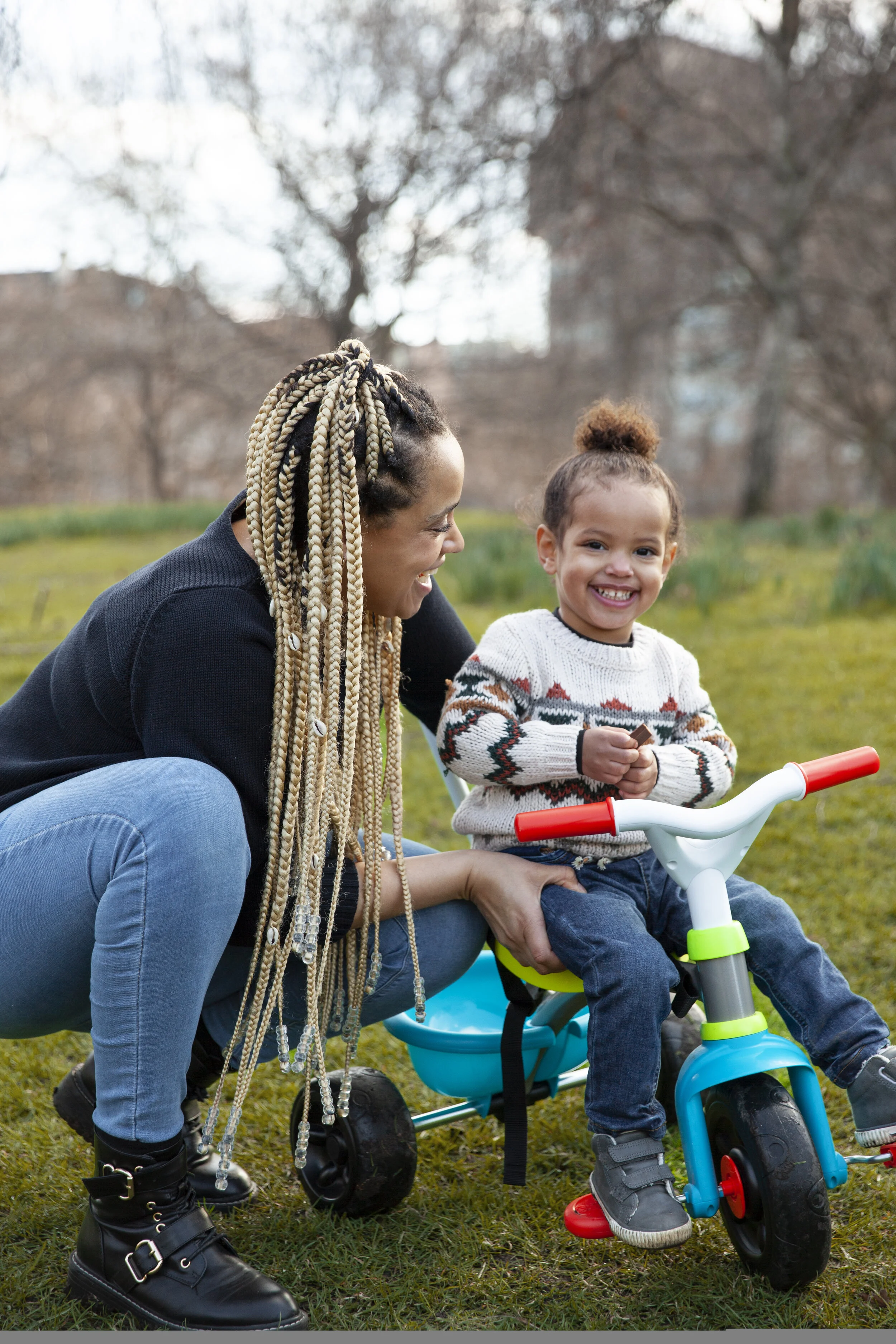 Woman and young girl outside on grassy area, girl sitting on balance bike, woman supporting her, trees and buildings in background.