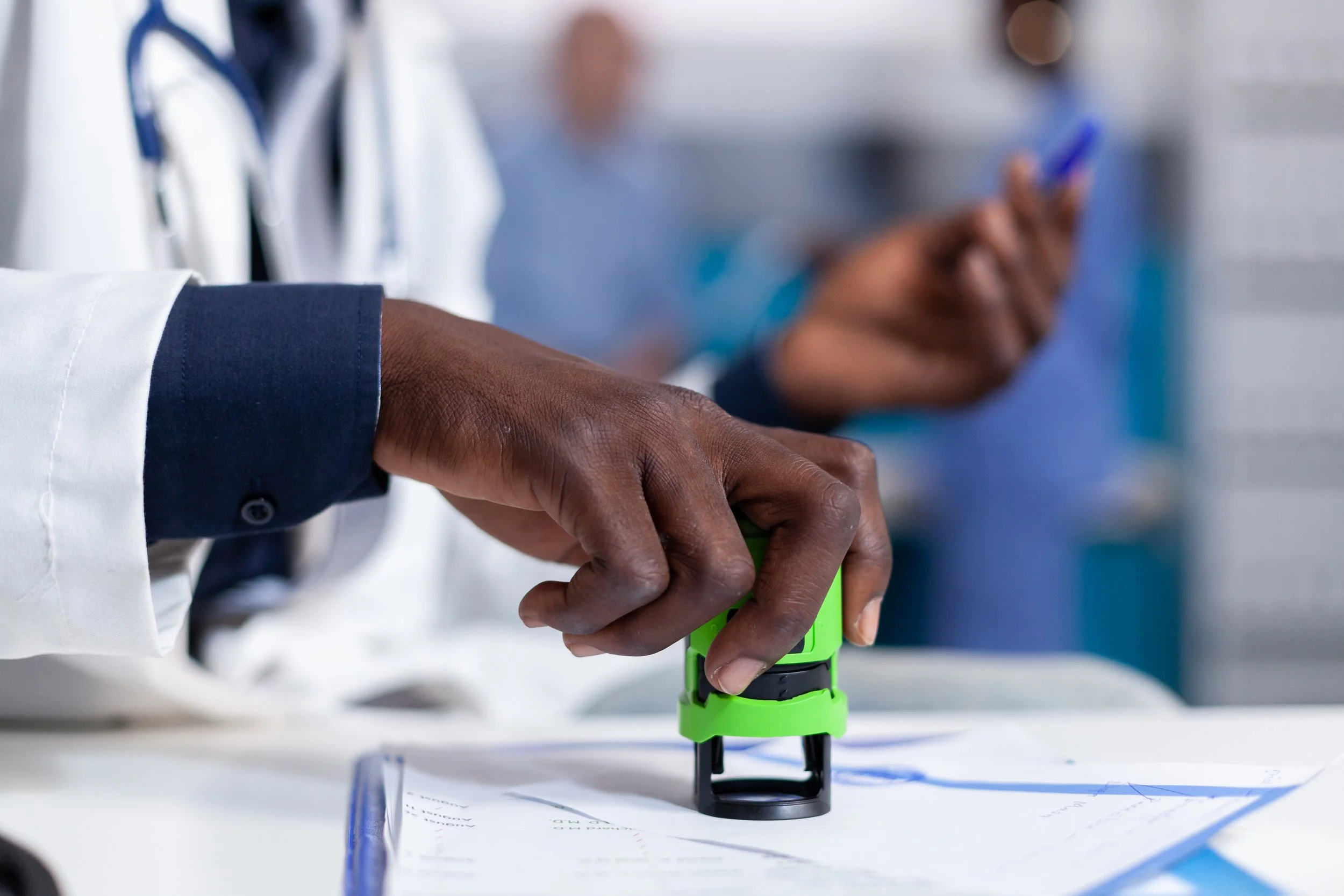 Close-up of a person's hand pressing a stamp on a document, with medical professionals in the background.