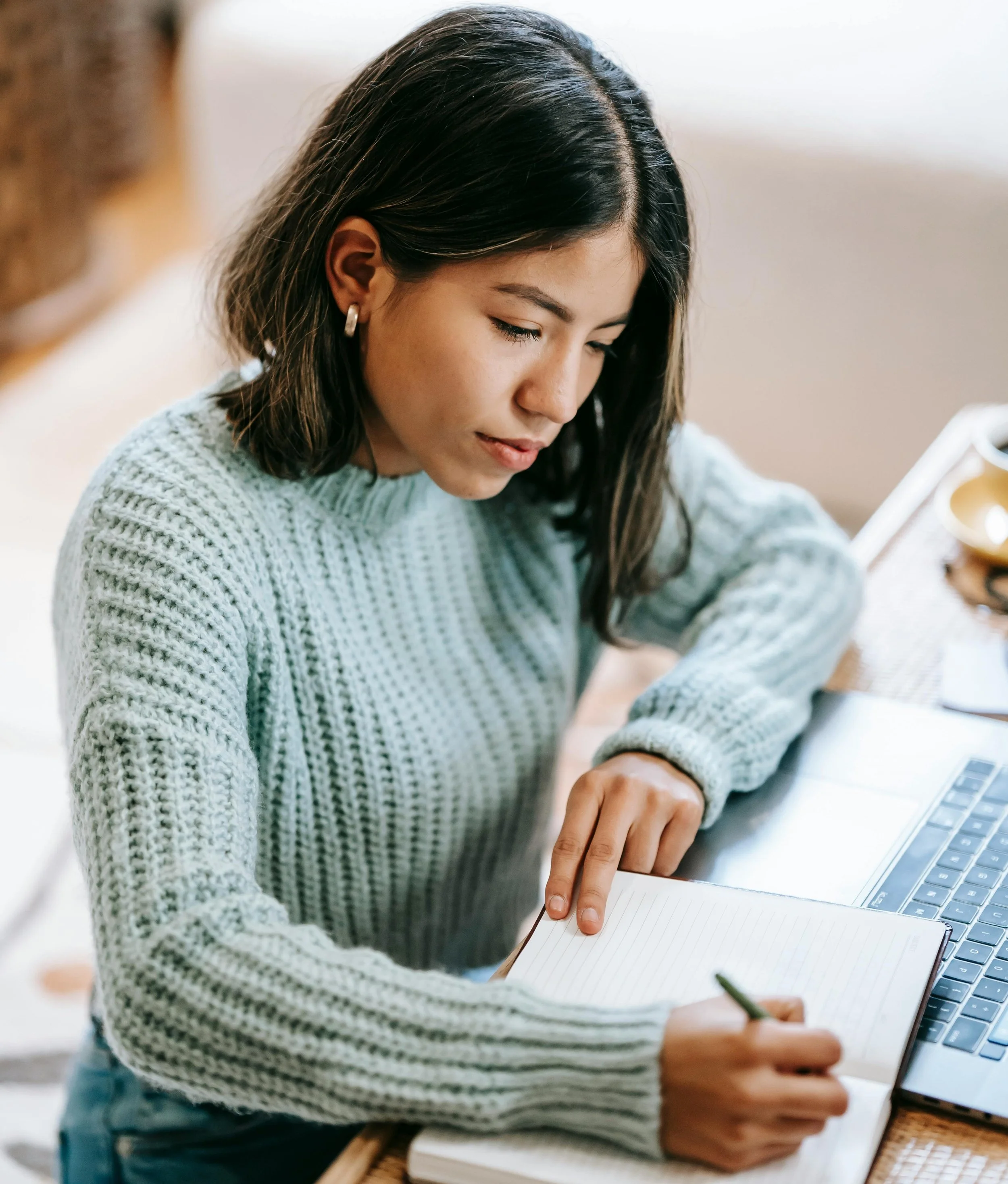 A young woman in a gray sweater writing in a notebook with a pen while working on a laptop at a desk.