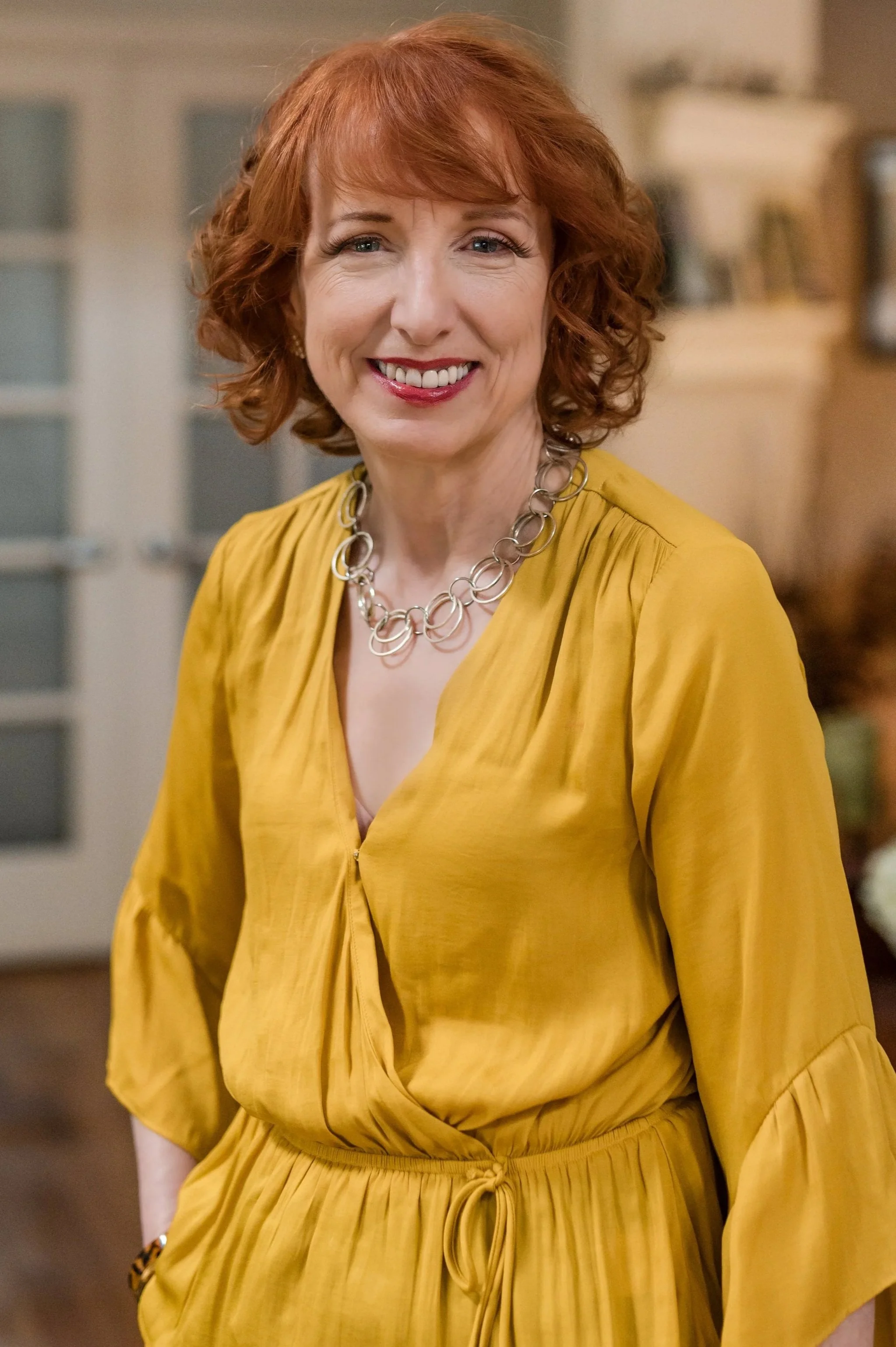A woman with red hair wears a mustard-yellow dress with a necklace and smiles in an indoor setting.