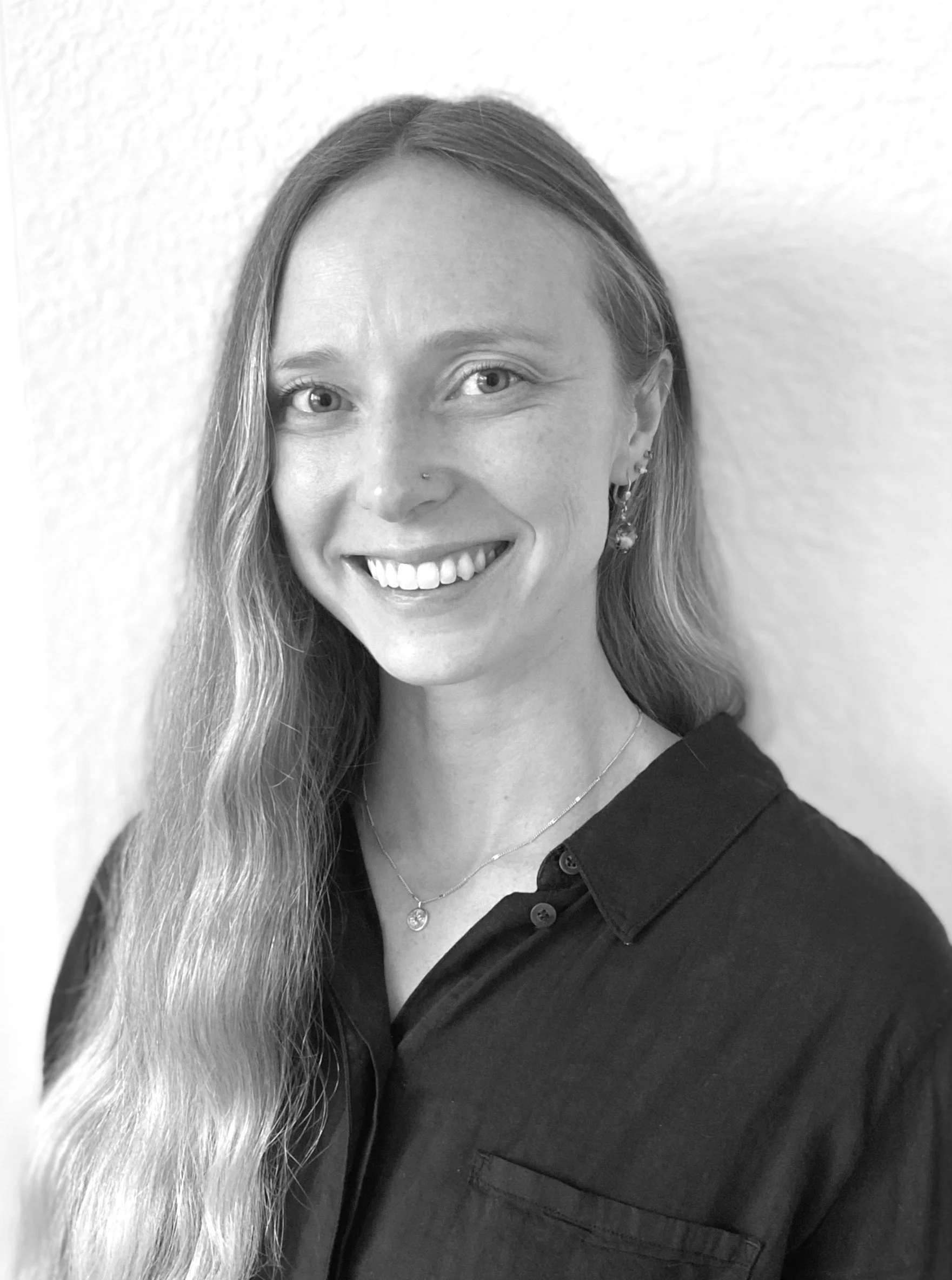 Black and white portrait of a smiling woman with long, wavy hair, wearing earrings and a necklace, standing against a plain wall.