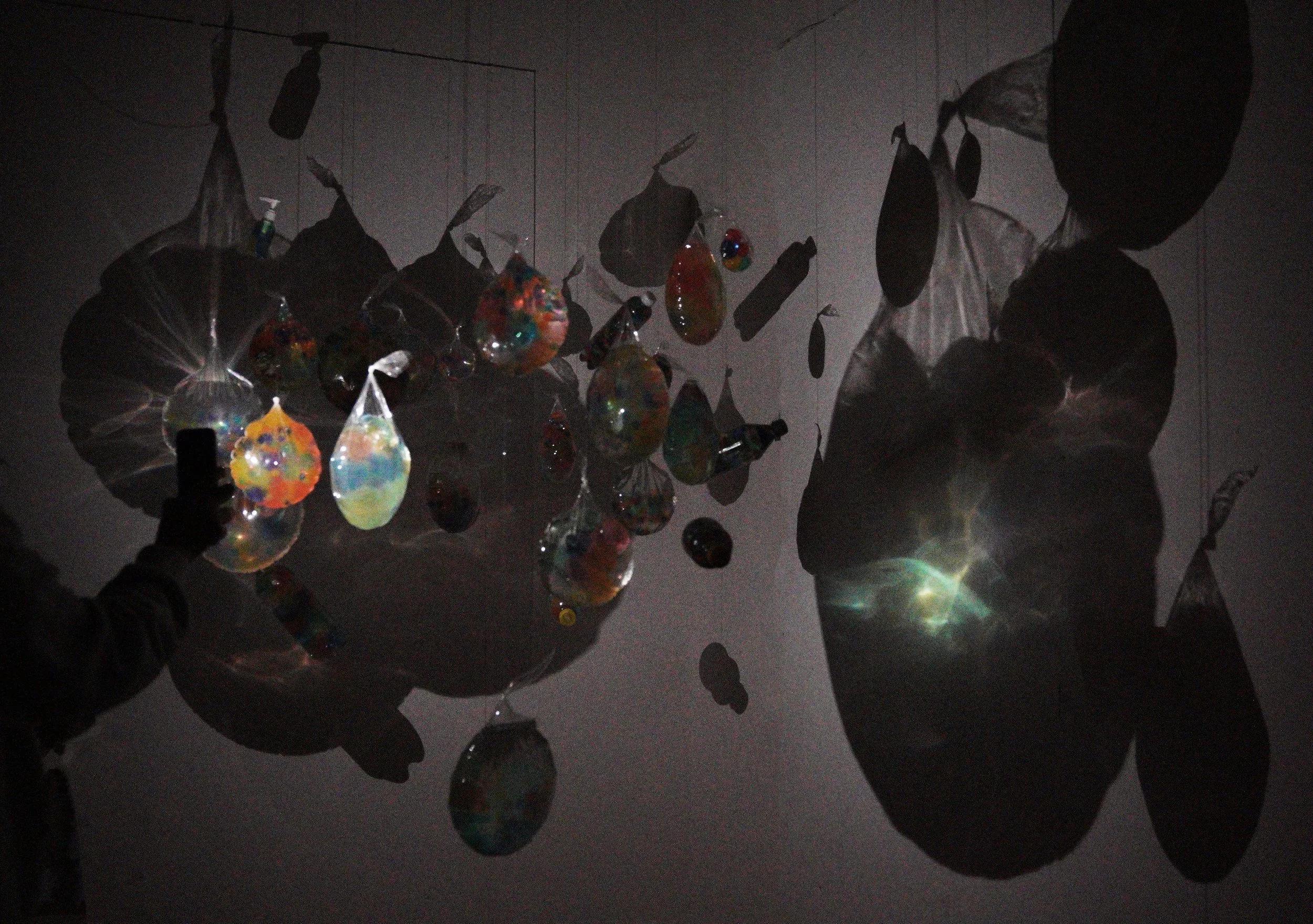 An art installation featuring hanging glass orbs and leaf-shaped objects with dramatic shadows cast on the wall.