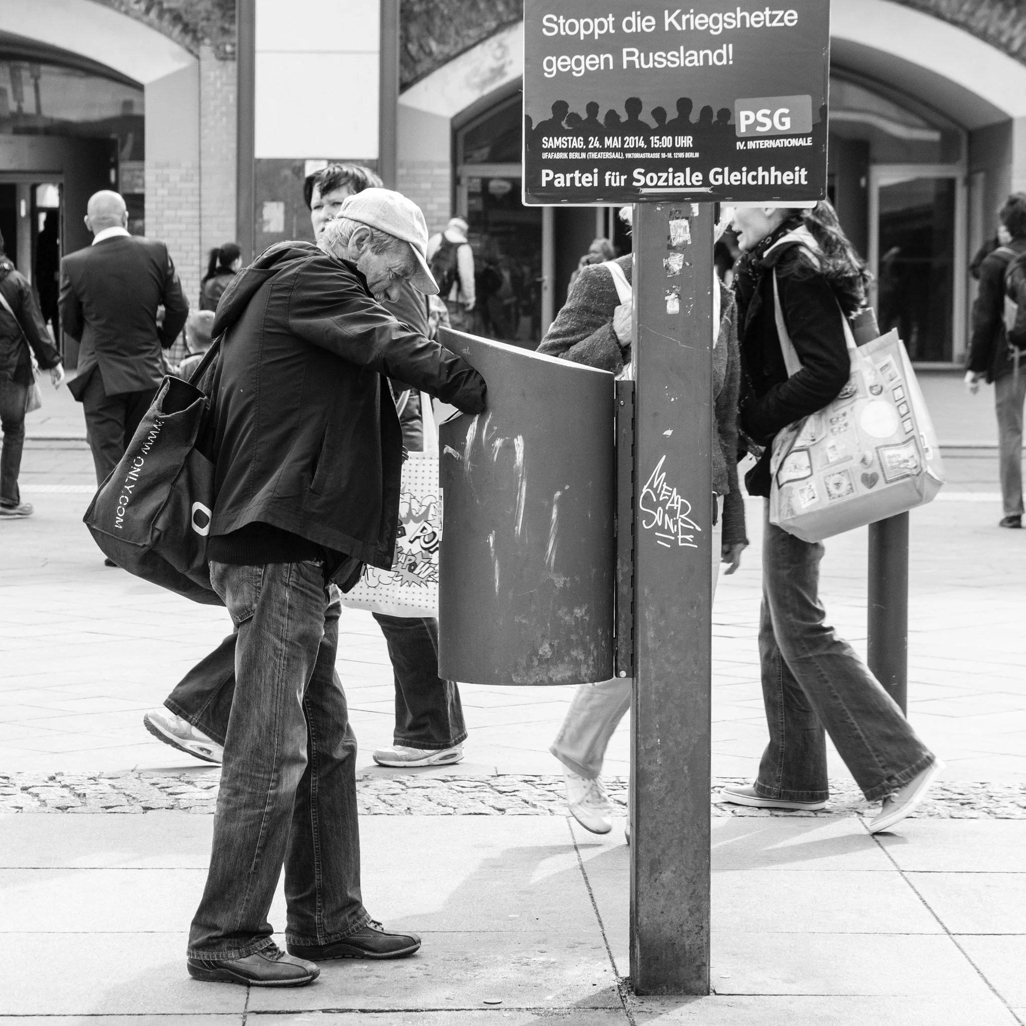Black and white photo of a man leaning over a donation or help box on a city street, surrounded by pedestrians.