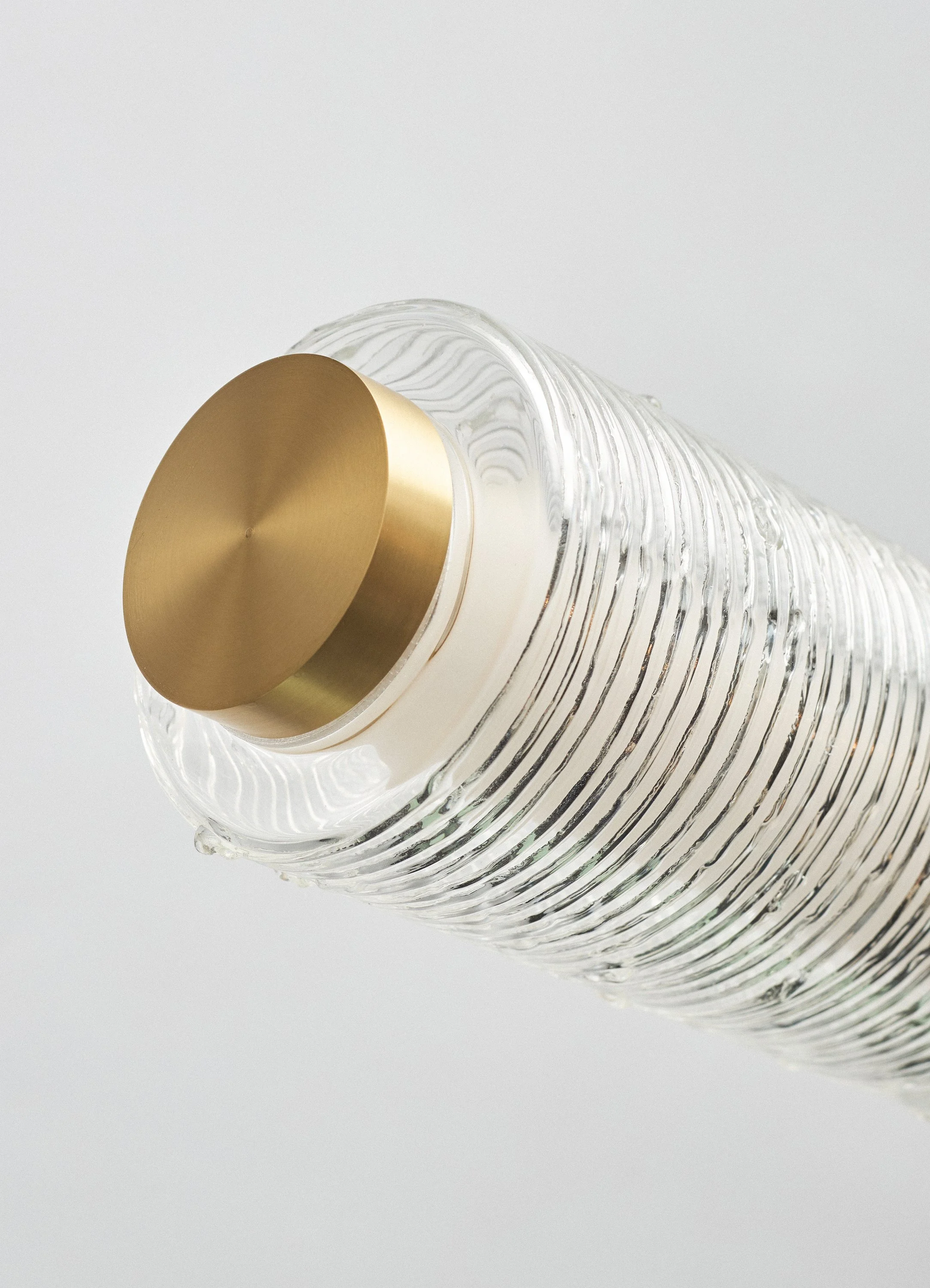 Close-up of a clear, ribbed glass bottle with a gold-colored, matte-finish cap, set against a plain light background.