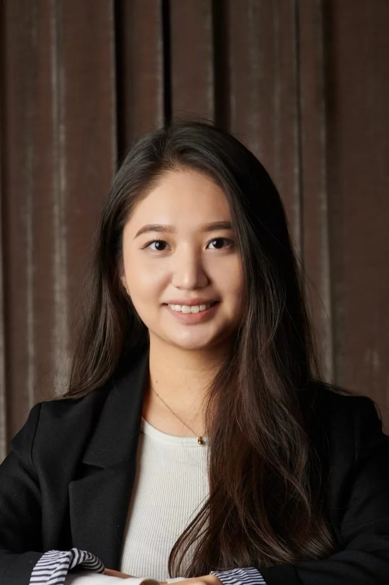 A young woman with long dark hair, smiling, wearing a black blazer and white top, sitting in front of a wooden background.