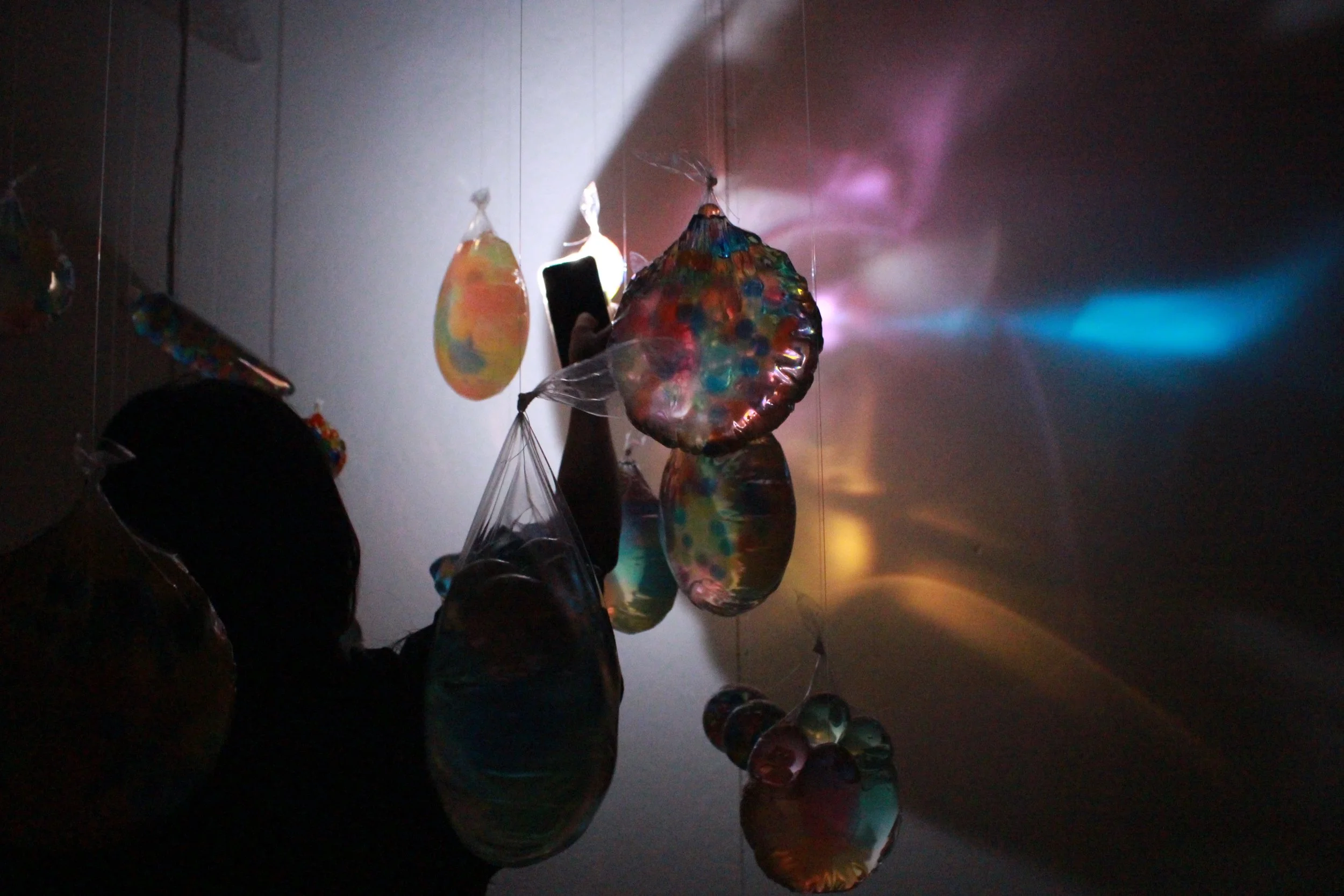 Colorful glass art sculptures hanging from ceiling, illuminated by a light source creating reflections and shadows on the wall.