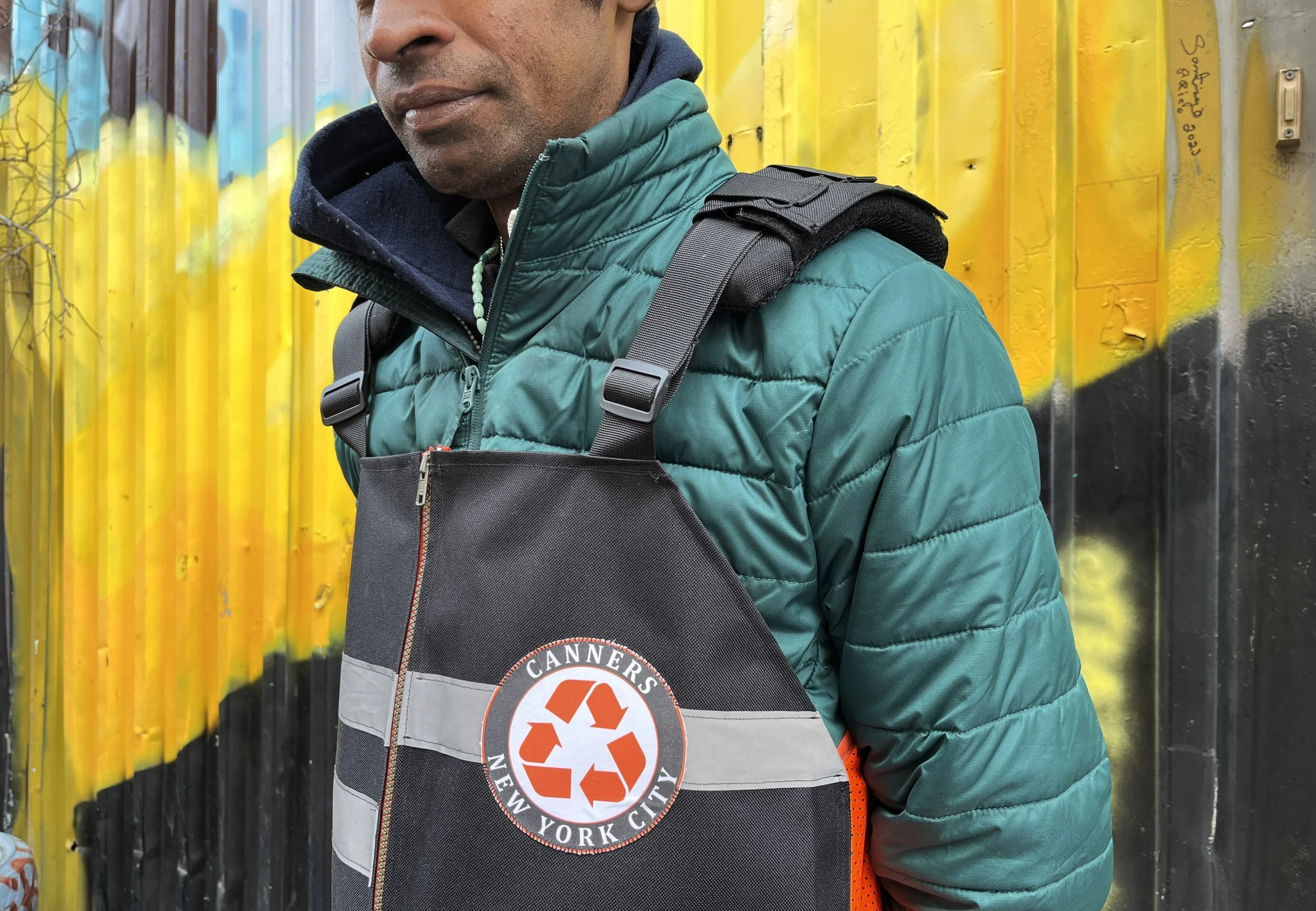 A person wearing a teal puffer jacket and a reflective vest with a recycling symbol and the words 'Canners New York City' printed on it, standing next to a yellow and black graffiti wall.