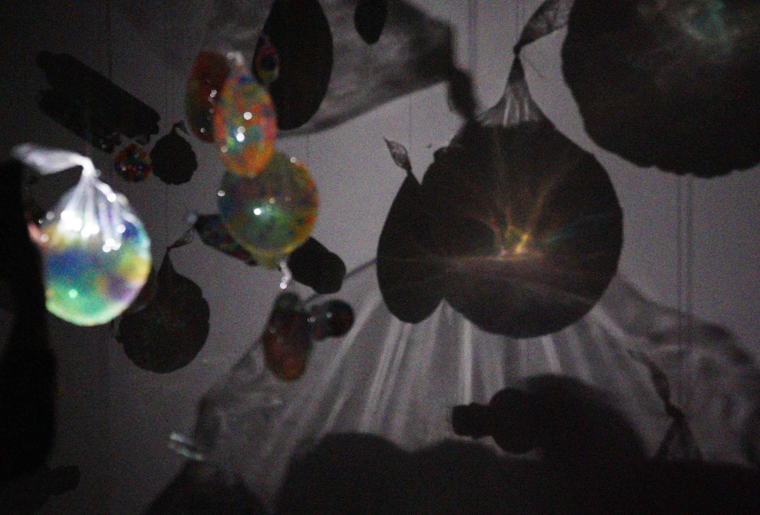 Colorful, shiny balloons hanging from a ceiling with dark leaves and a light source creating a starburst effect.