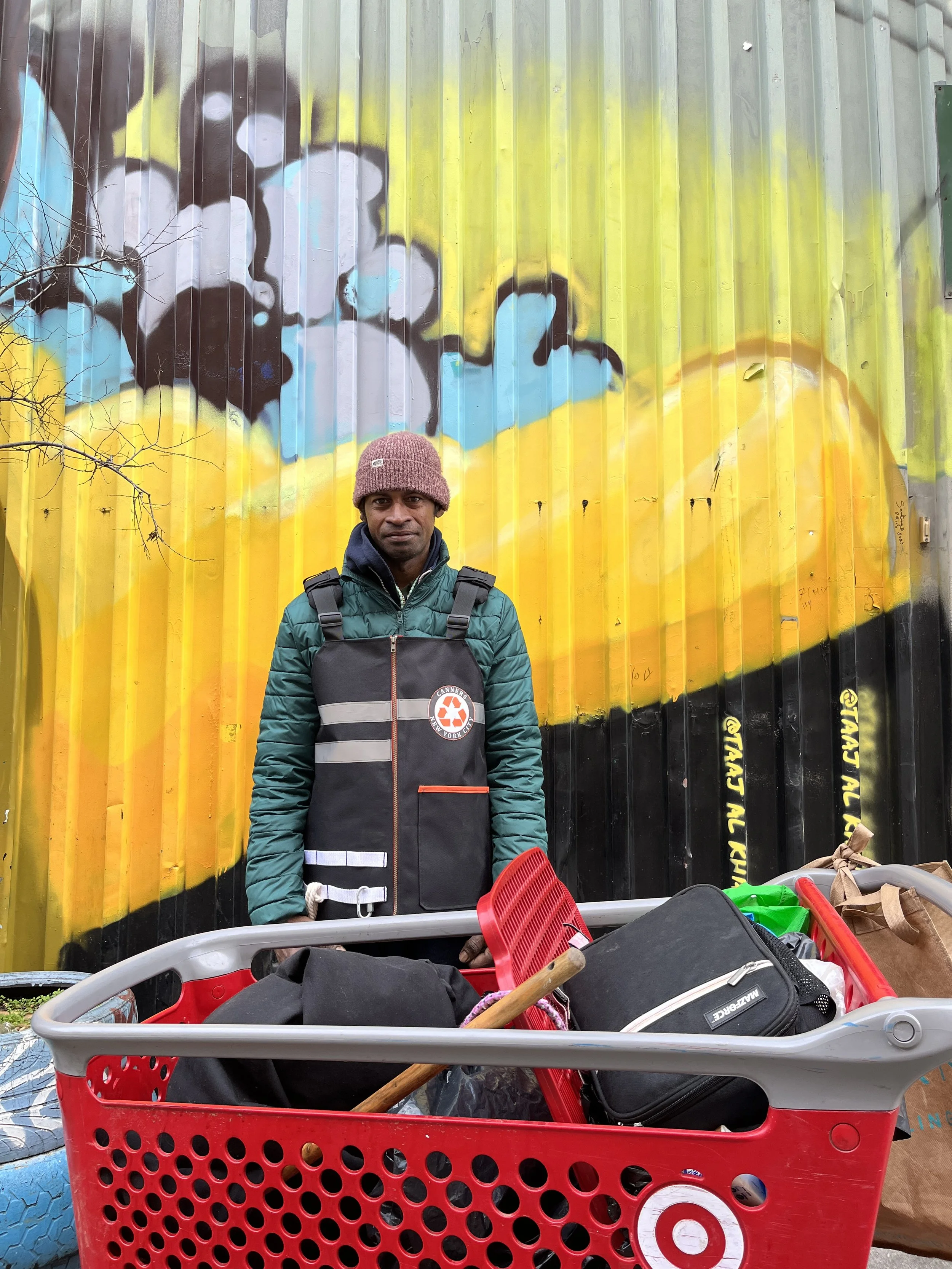 A man standing next to a shopping cart filled with various items including bags and a gardening tool, in front of a colorful graffiti mural on a metal wall.