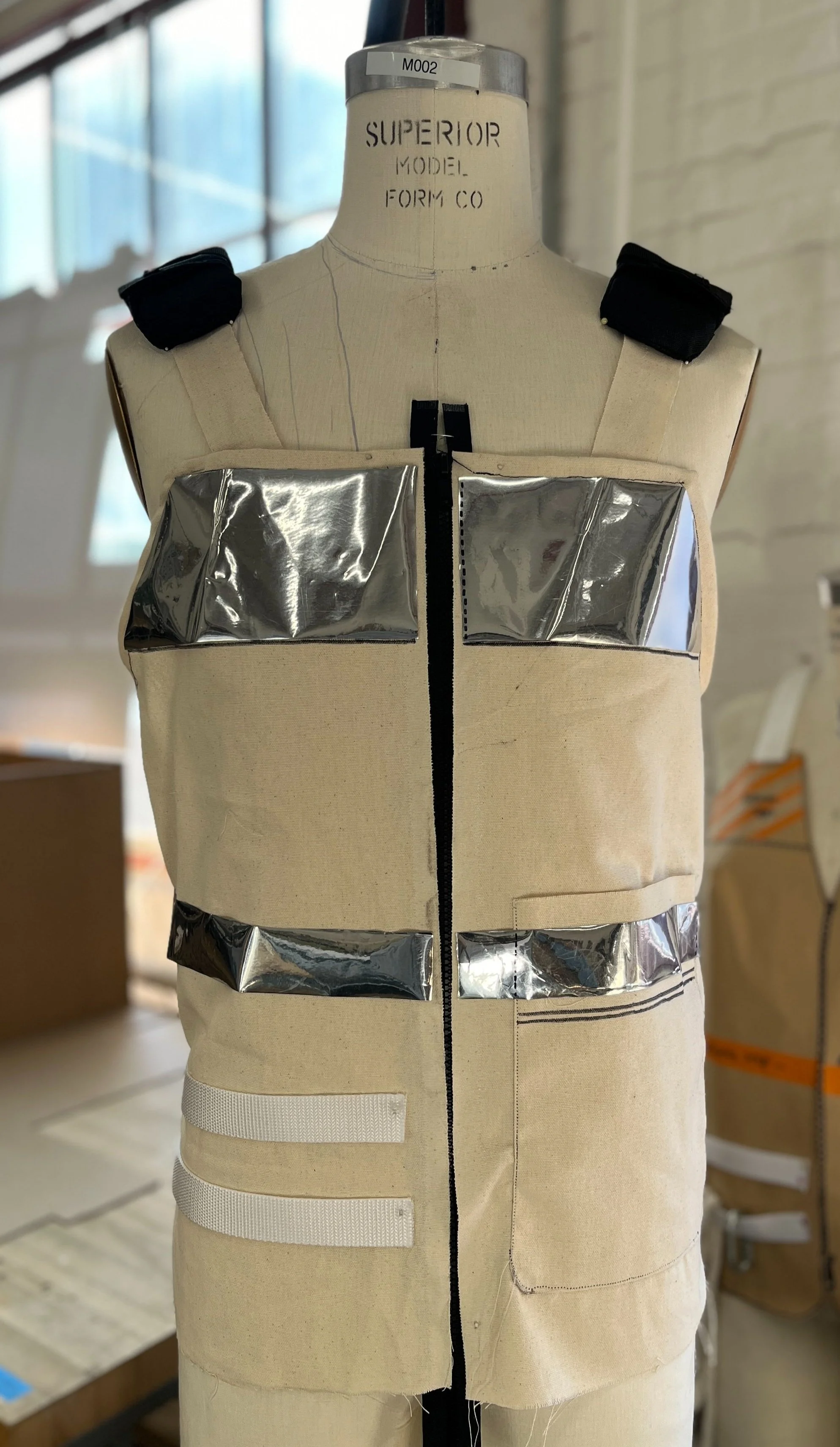 A dress form mannequin wearing a beige vest with reflective silver strips and black shoulder patches in a workshop setting.