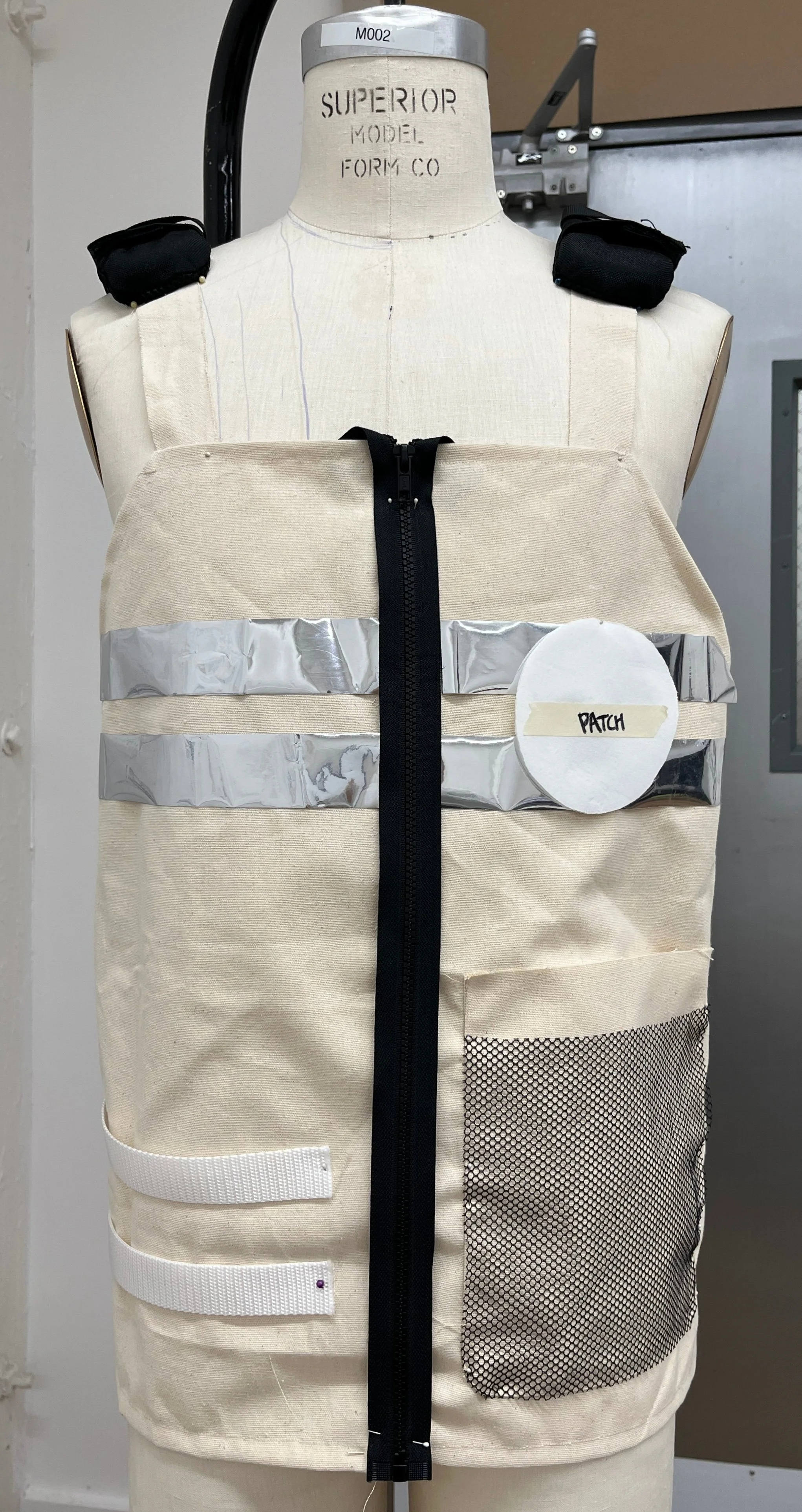 A dress form mannequin wearing an unfinished beige fabric vest with black shoulder pads, a central black zipper, two white straps, a round white patch labeled 'PATCH,' black mesh pocket, and horizontal silver reflective tape.